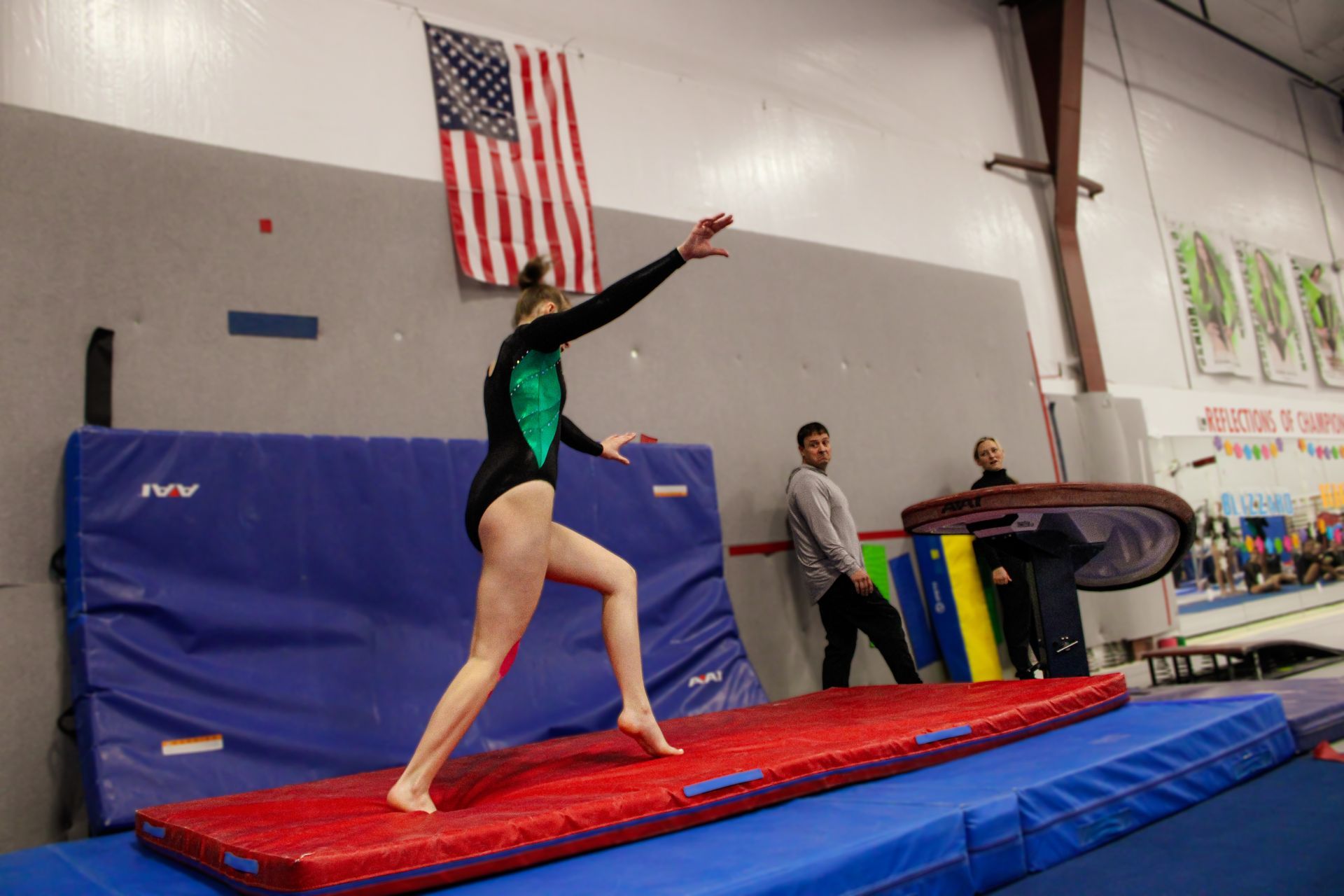 Gymnast on a red and blue mat mid-leap in a gym, with an American flag and coach nearby