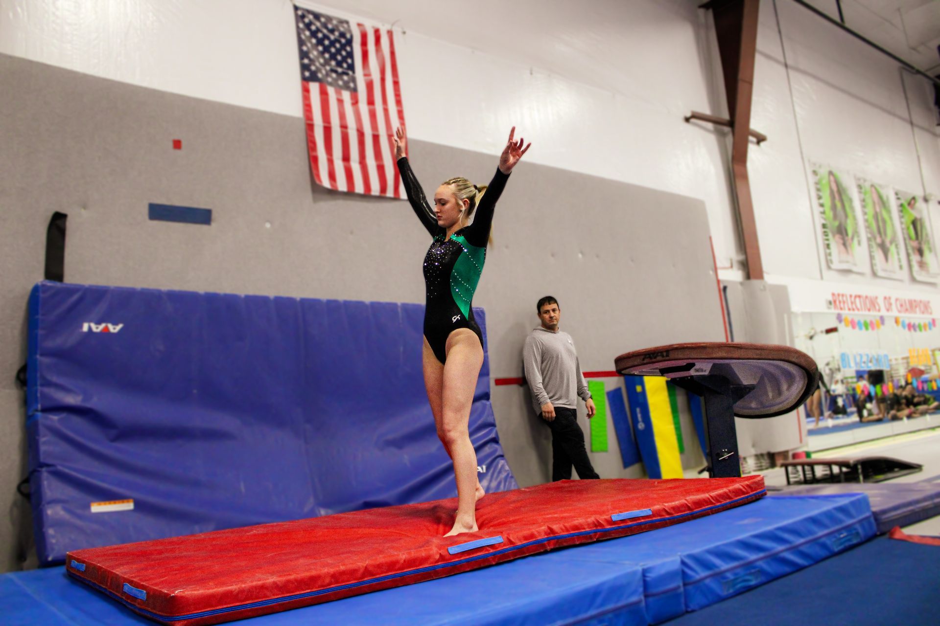 Gymnast balancing on a red beam mat in a gym, arms raised, with an American flag in the background
