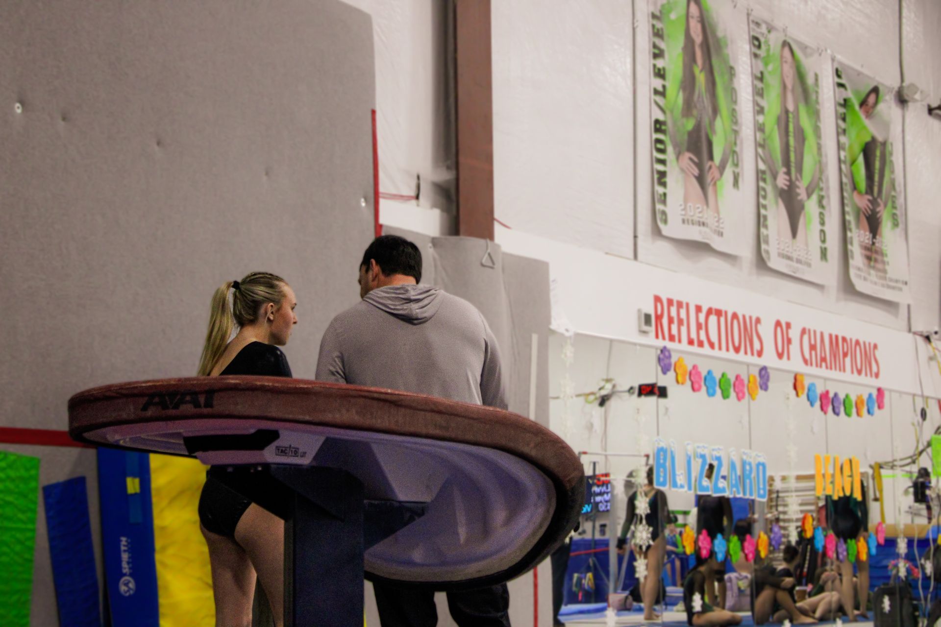 Two people stand near a gym floor with athletes and a banner in the background.