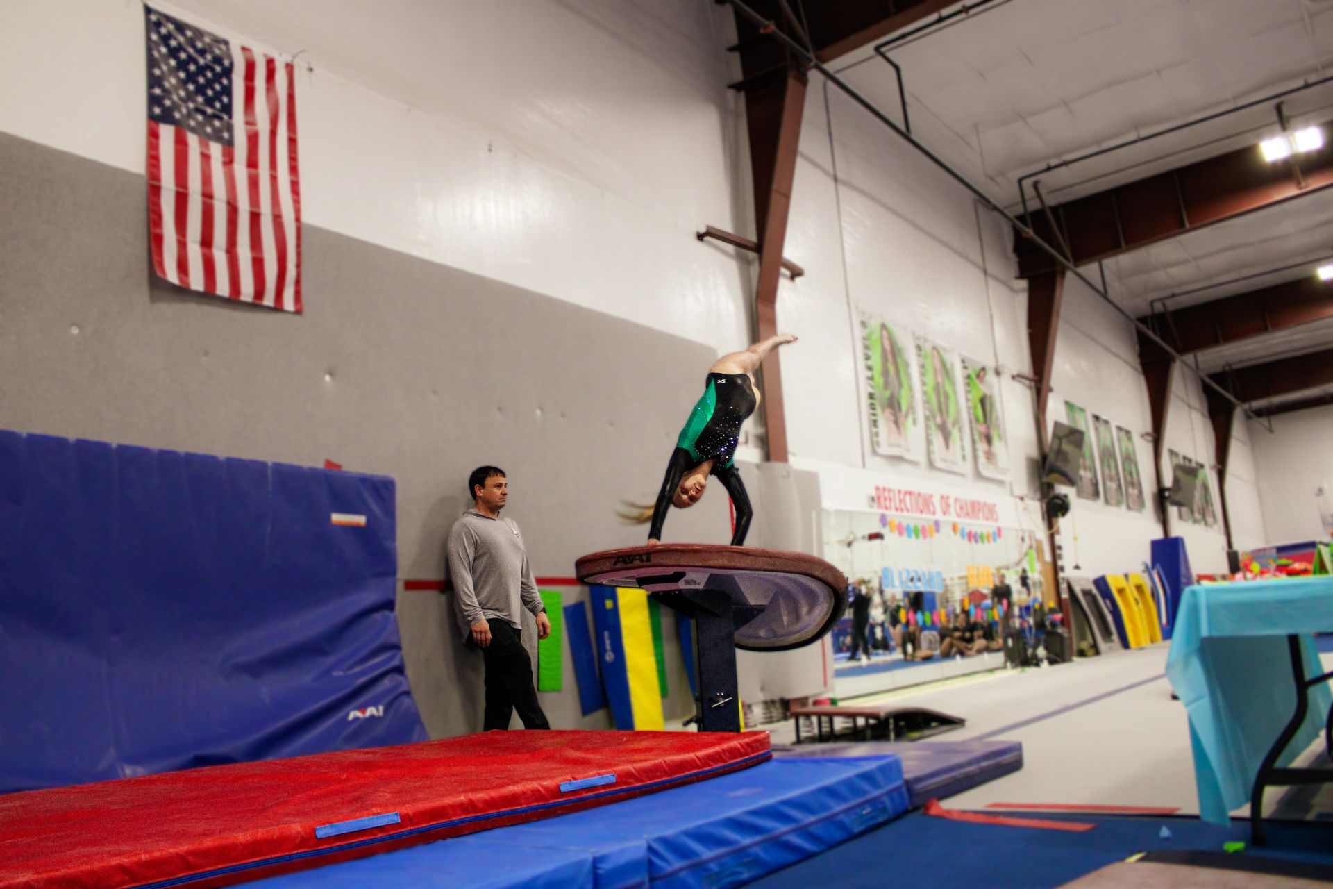 Gymnast flipping on a springboard in a gym, with mats and a coach nearby