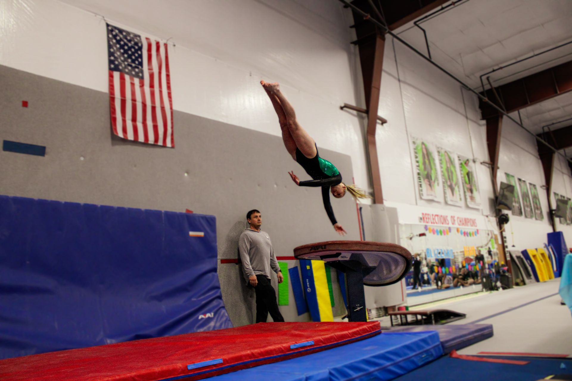 Gymnast flipping over a trampoline in an indoor training gym, with a coach nearby and mats below.