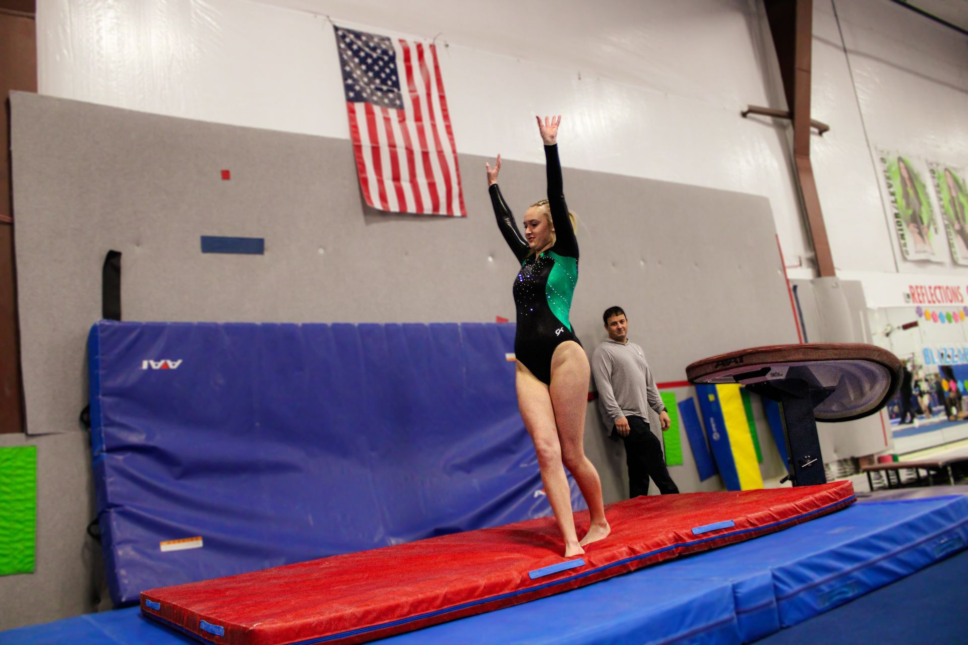 Gymnast on a red mat raises one arm in a gym, with an American flag on the wall.