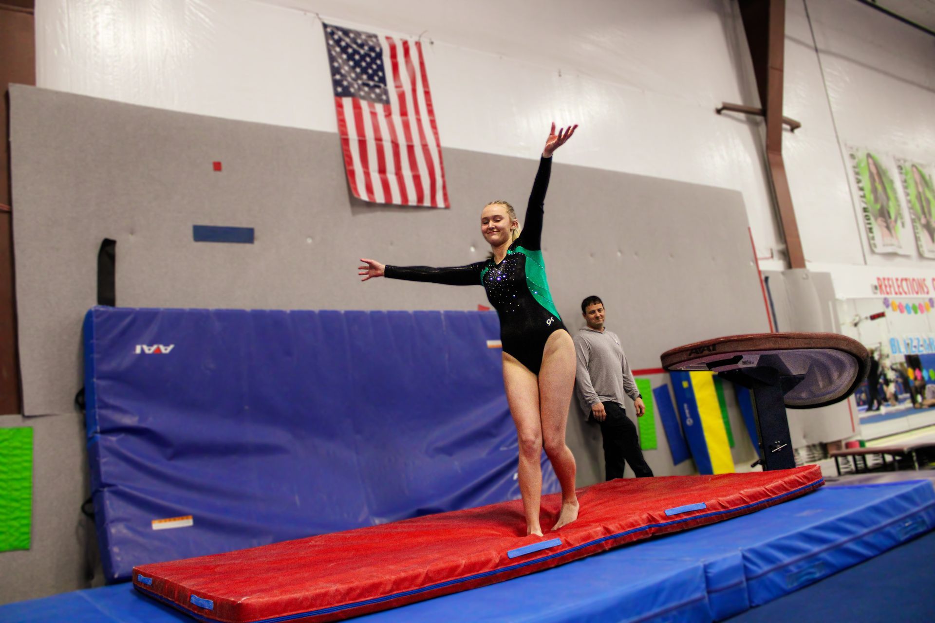 Gymnast balancing on a red runway mat inside a gym, arms raised beneath an American flag.