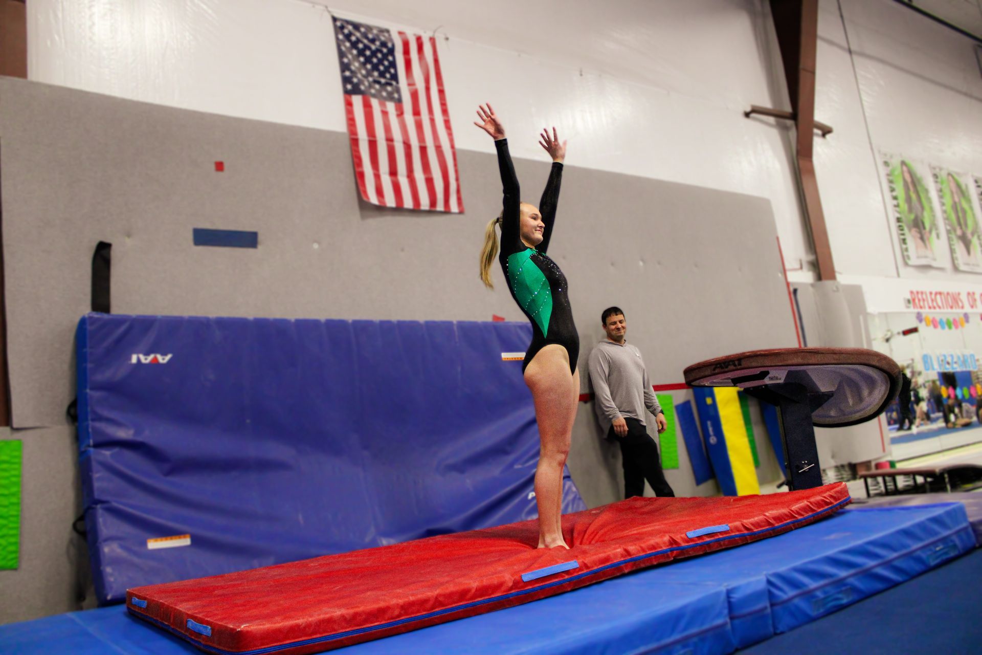 Gymnast on red mat with arms raised in a gym, with coach nearby and an American flag on the wall