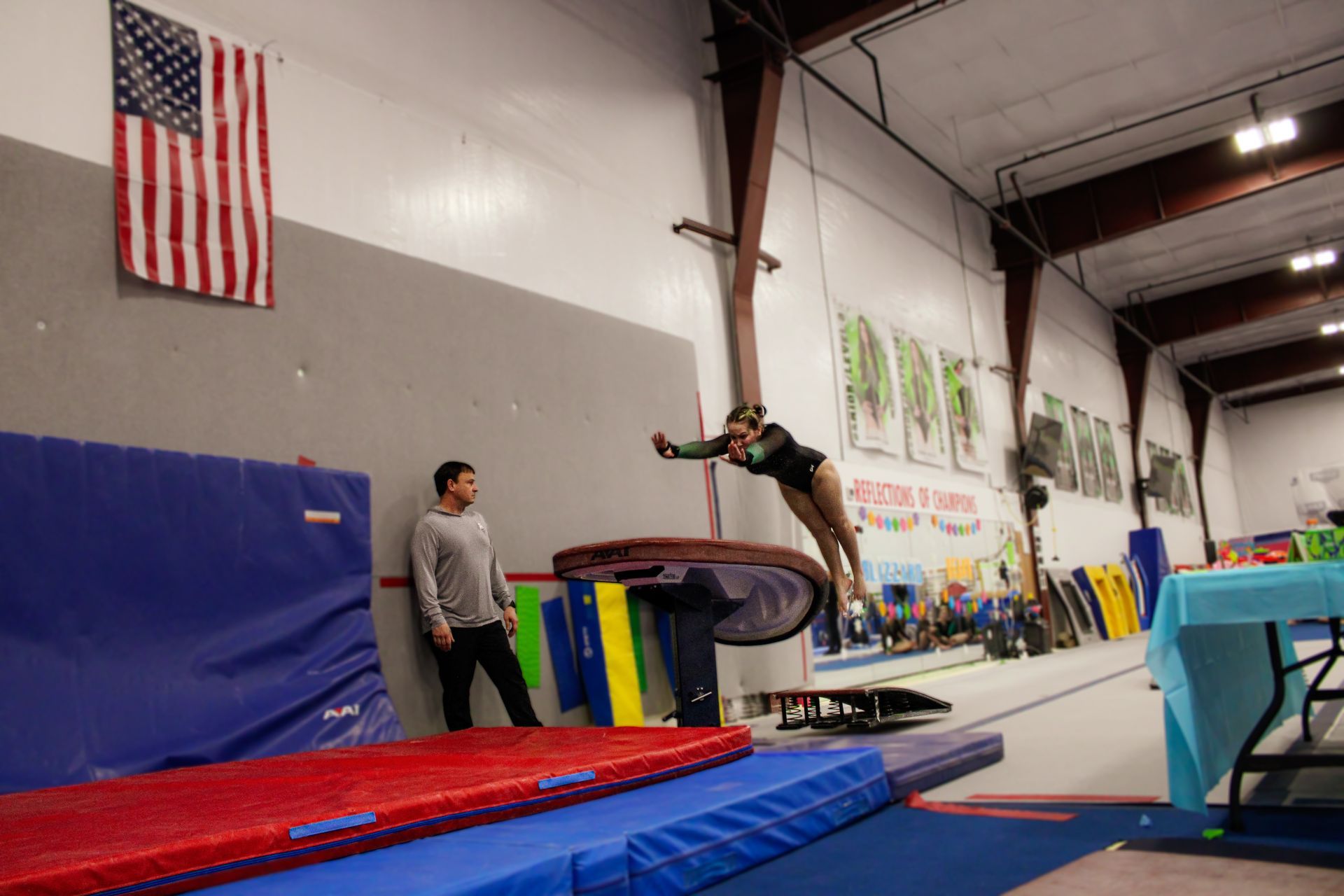 Gymnast vaulting over a trampoline in an indoor gym, with a coach nearby and an American flag on the wall