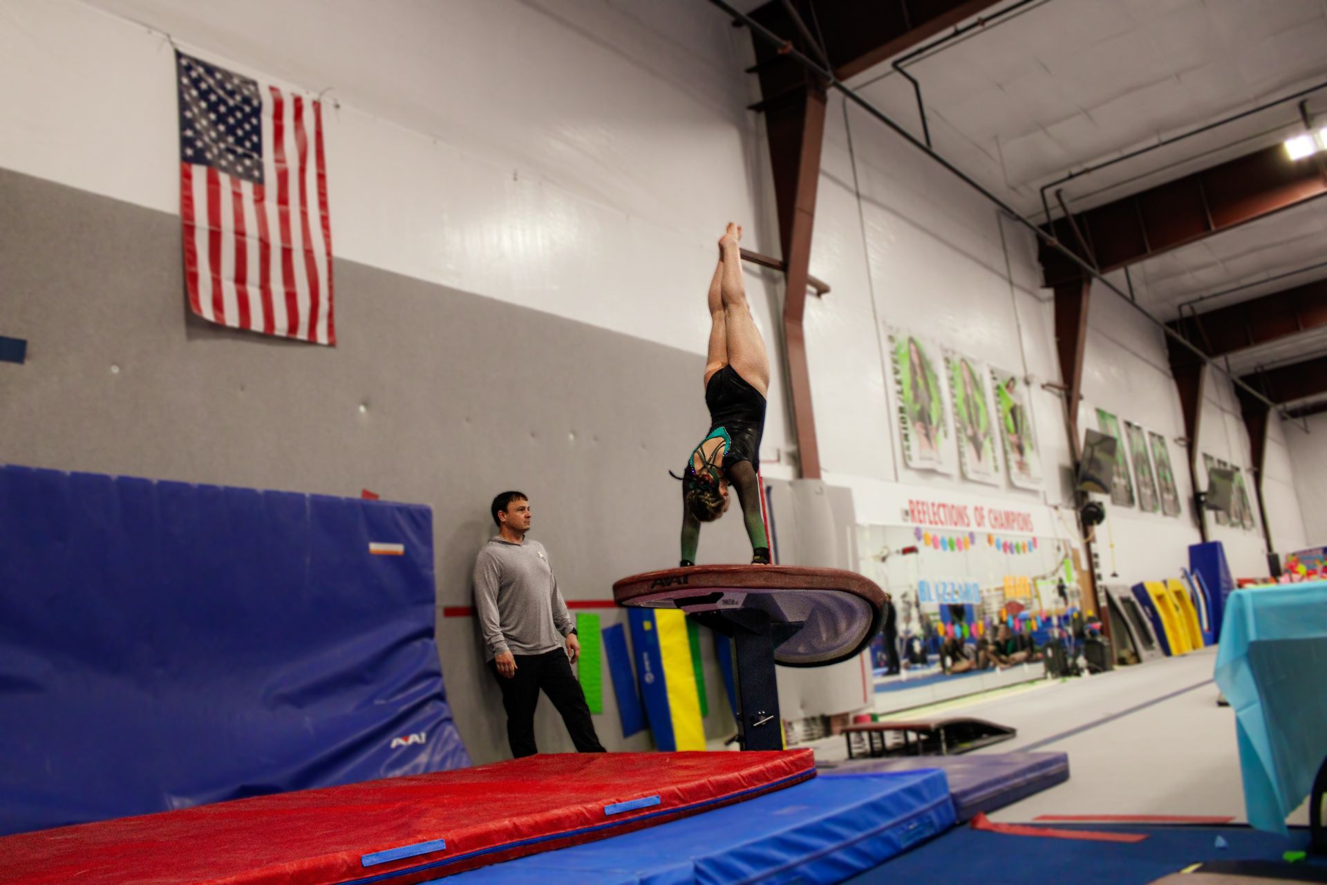 Gymnast balancing upside down on a vault table in a gym, with a coach watching nearby.