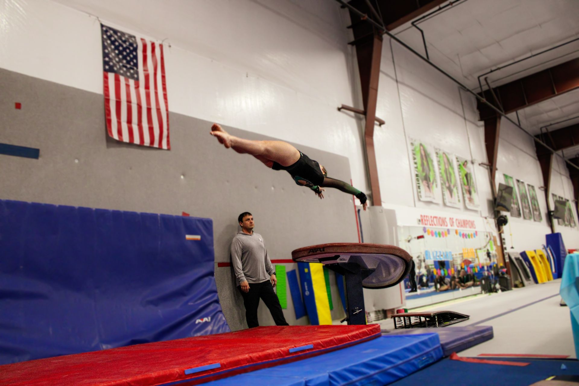 Gymnast mid-flip over a padded mat in an indoor gym, with a coach watching nearby.