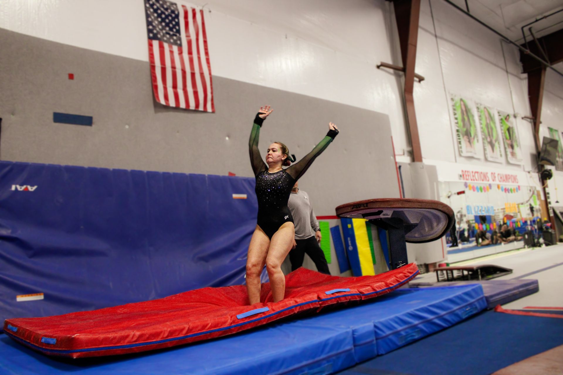Gymnast on a red mat with arms raised in a gym, beside blue crash mats and an American flag.