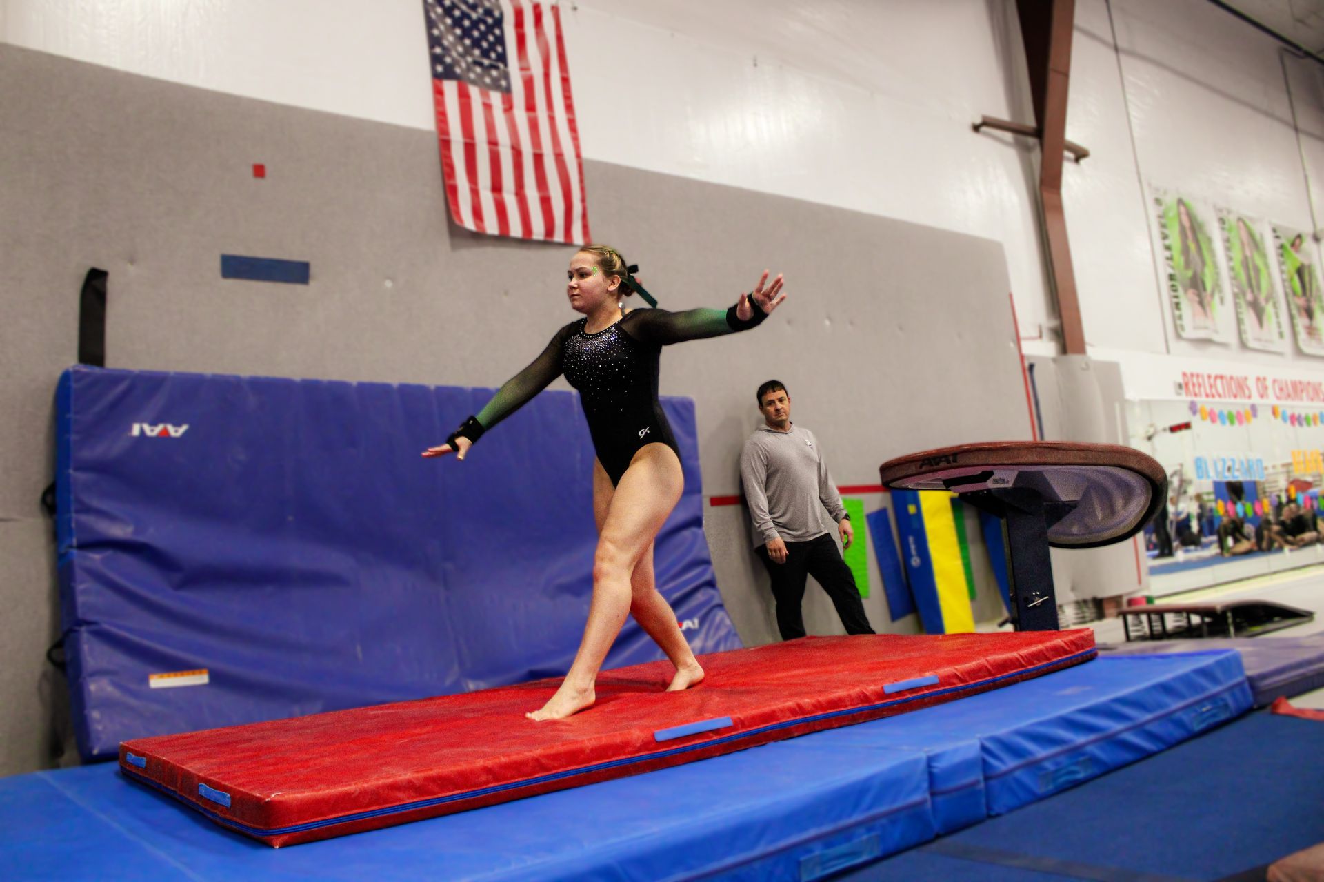 Gymnast balancing on a red mat in an indoor gym, with a coach nearby and an American flag above.