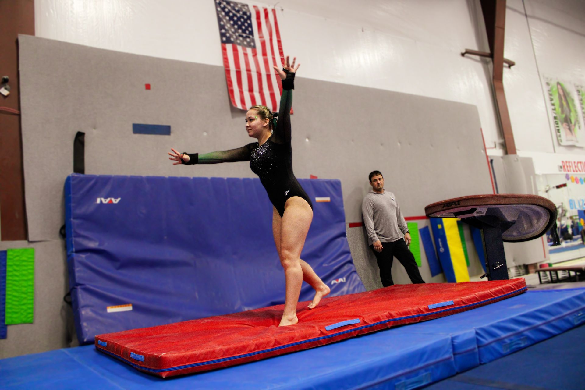 Gymnast balancing on a red mat in a gym, with an American flag and coach in the background