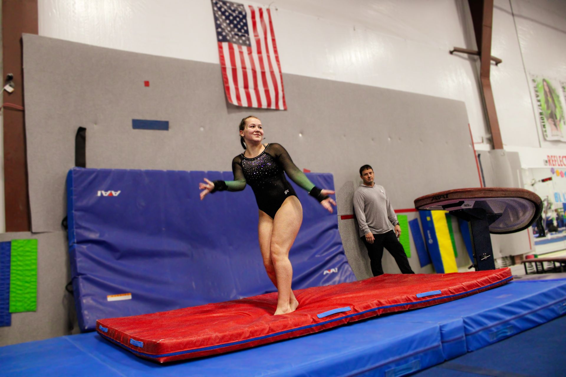 Gymnast balancing on a red beam in a training gym, with mats and an American flag in the background