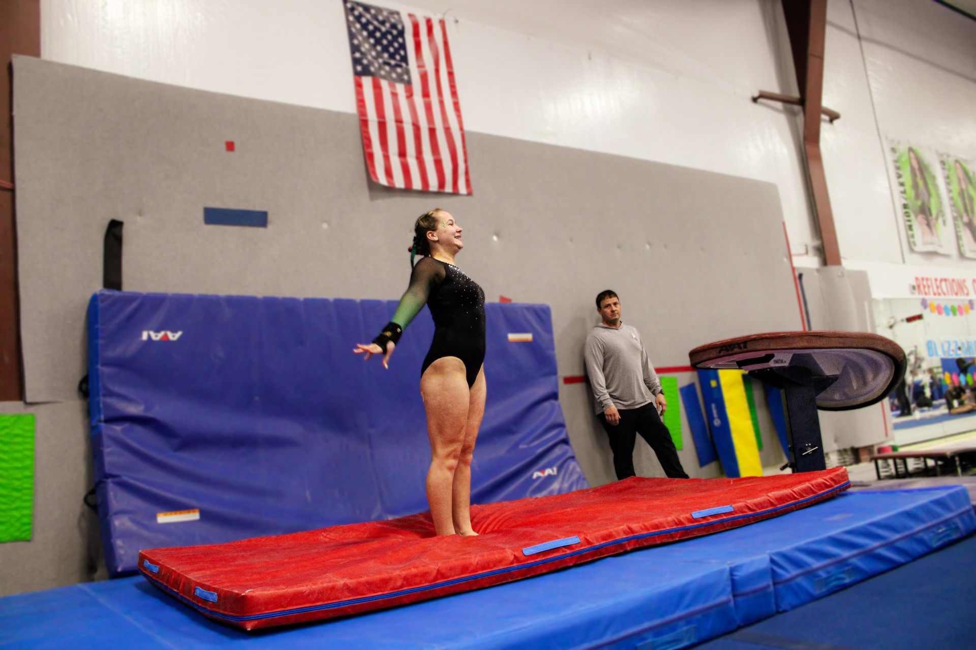 Gymnast standing on a red mat in a gym, with a coach nearby and an American flag on the wall.