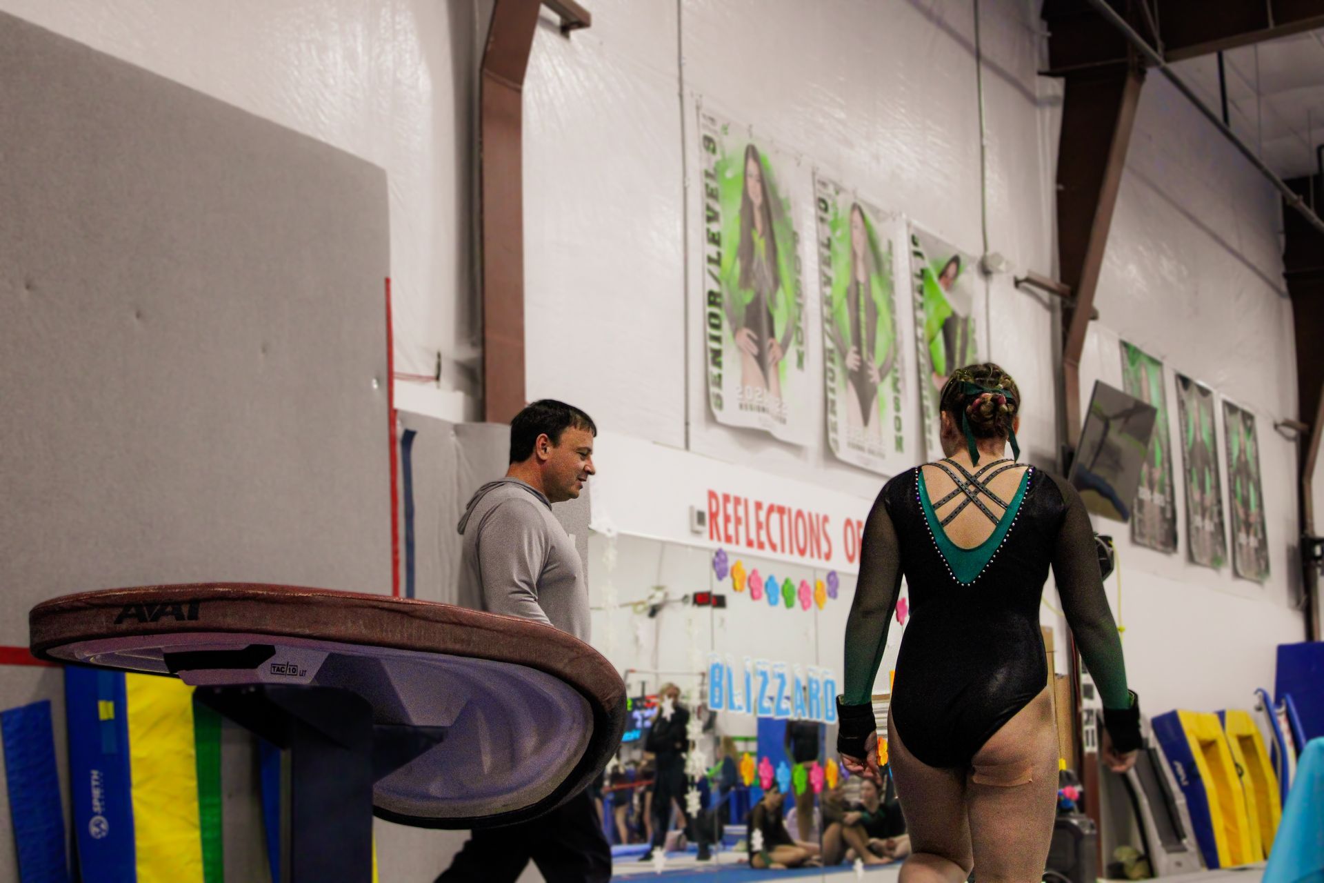 Indoor gymnastics gym with two people near a balance beam and wall-mounted posters