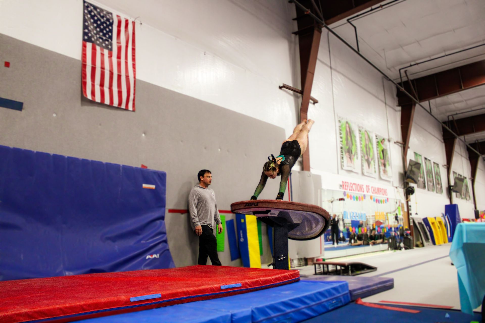Gymnast mid-air over stacked mats in an indoor training gym, with a coach nearby and an American flag on the wall