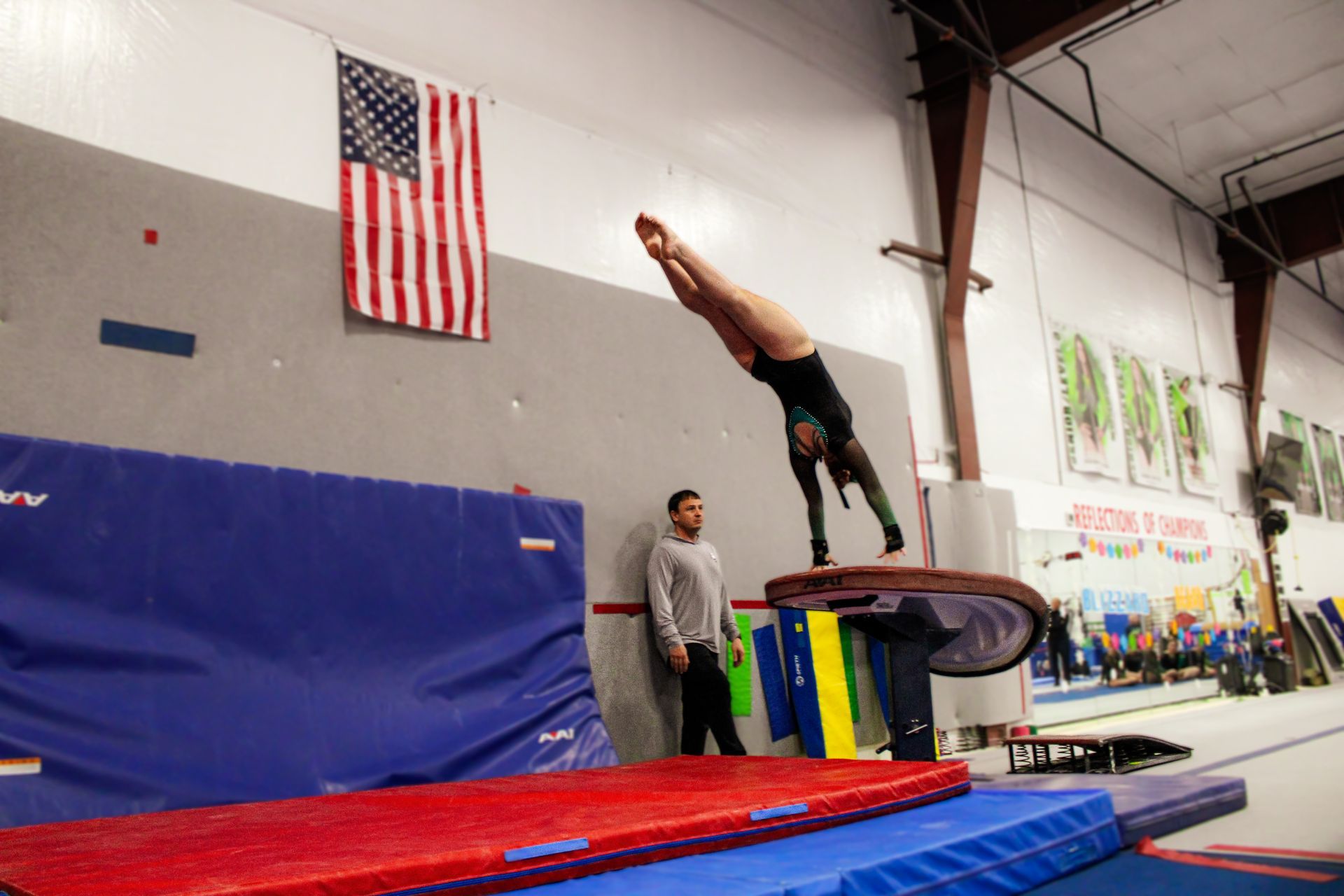 Gymnast performing a backflip above a foam pit in an indoor training gym, coach watching nearby.