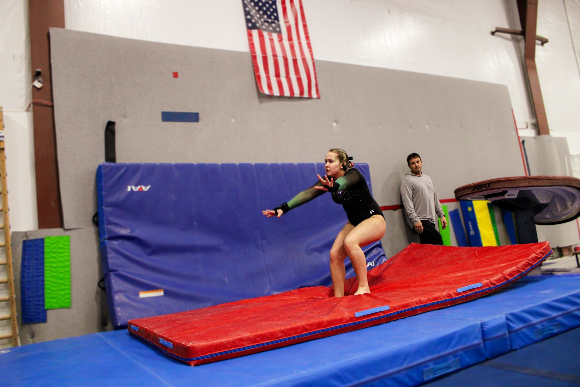 Gymnast balancing on a red mat in a gym, with blue crash mats and an American flag in the background