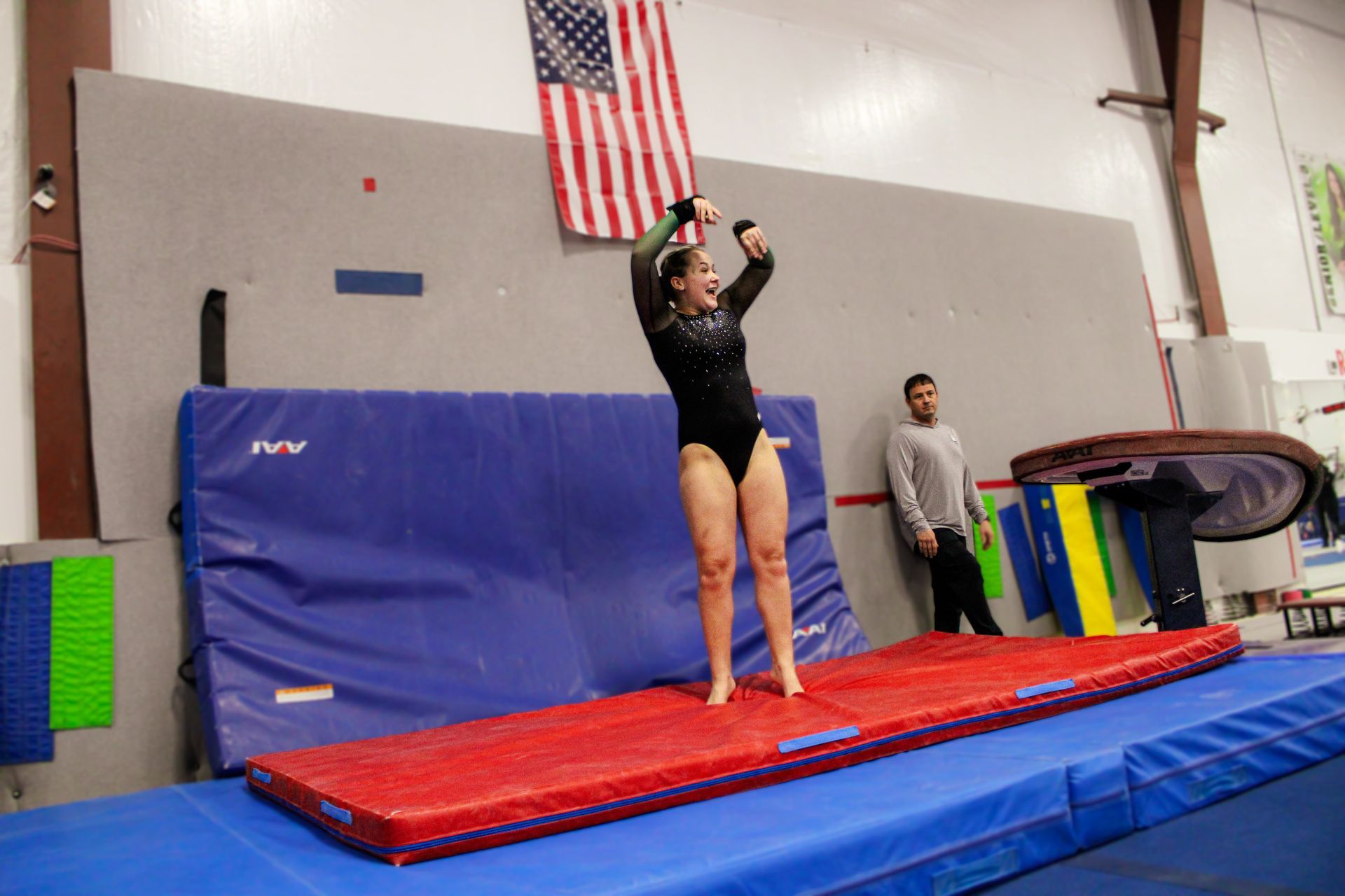 Gymnast on a red mat raising arms in a gymnasium with blue crash mats and an American flag.