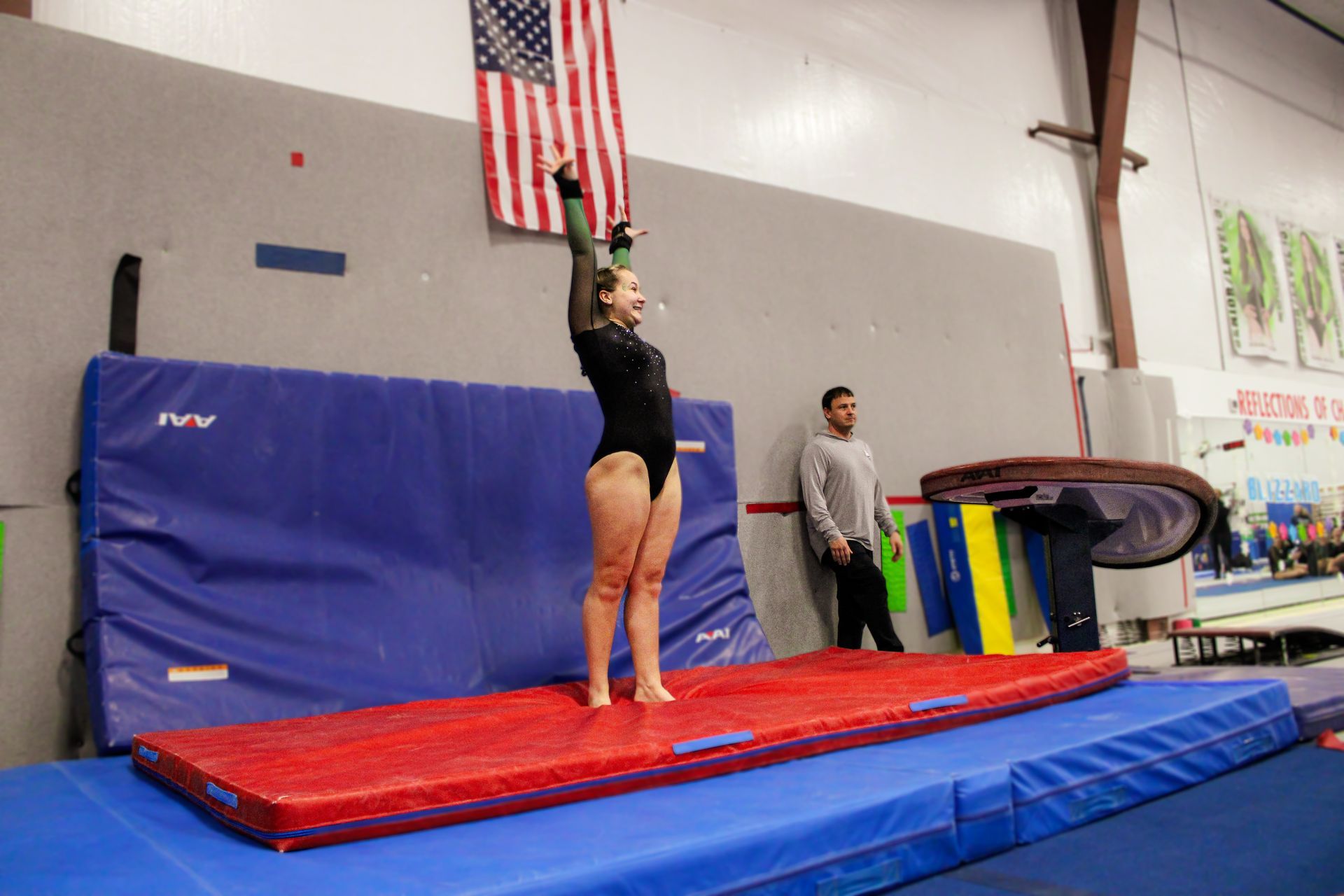 Gymnast balancing on hands on a red mat in a gym, with an American flag and coach nearby.