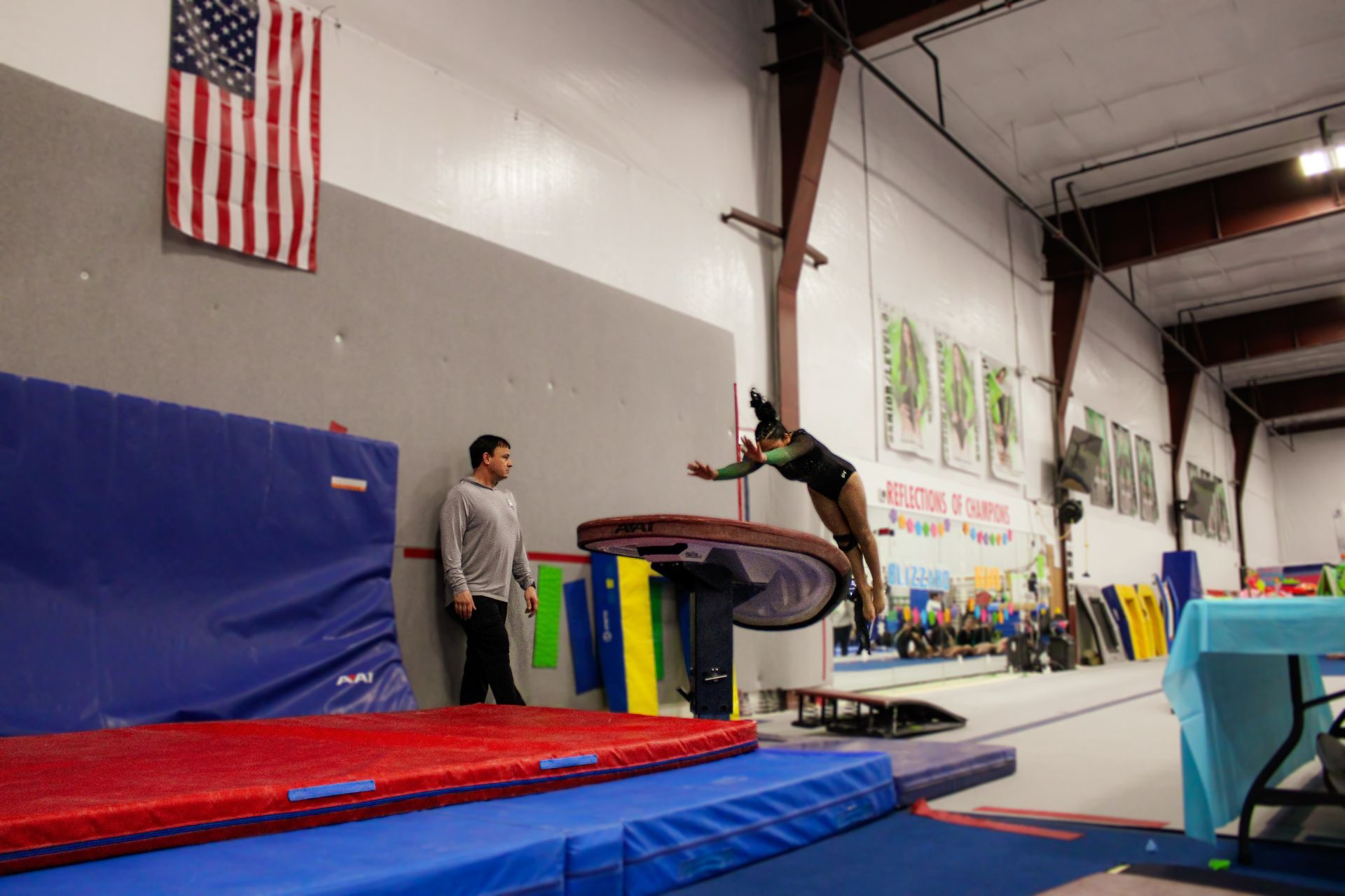 Gymnast mid-air over a vault in a gym, with a coach watching nearby.