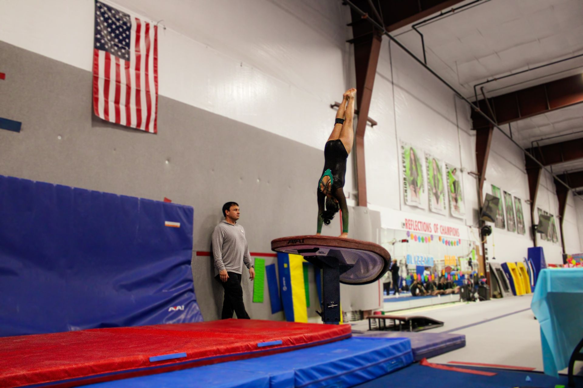 Gymnast hanging upside down on a bar in an indoor training gym with mats and an American flag.