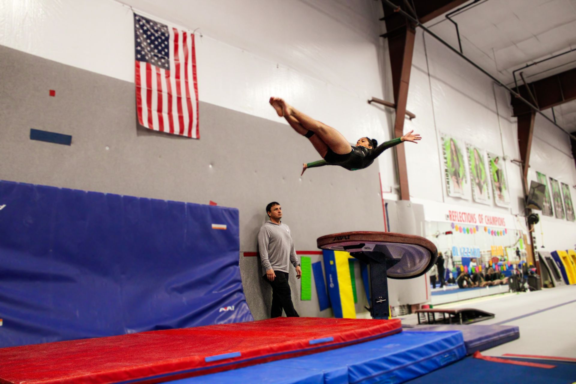 Gymnast flipping over a red-and-blue mat in an indoor training gym, with an American flag on the wall.