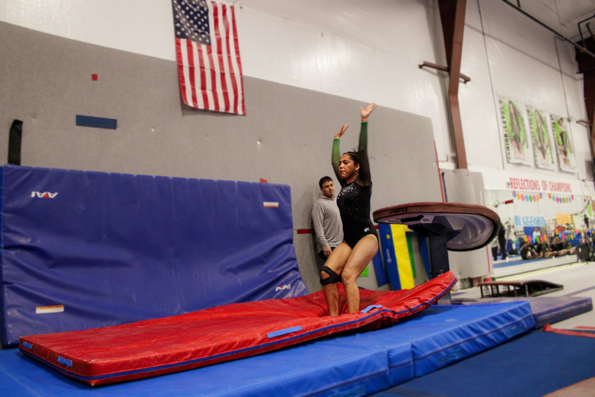 Gymnast balancing on a red beam in a gym, with a coach spotting nearby and mats below