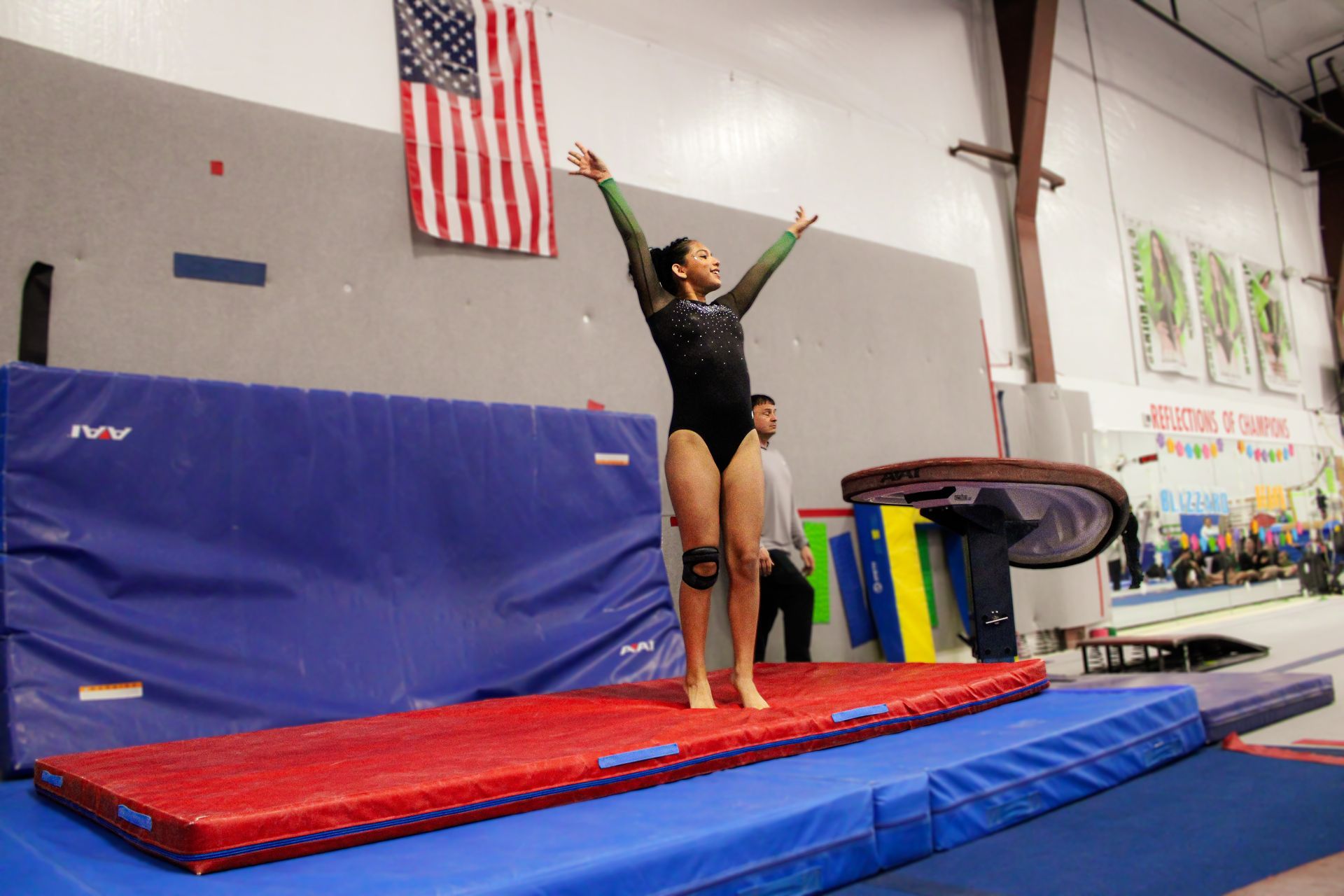 Gymnast mid-air on a red mat in a gym, with arms raised and an American flag hanging behind.
