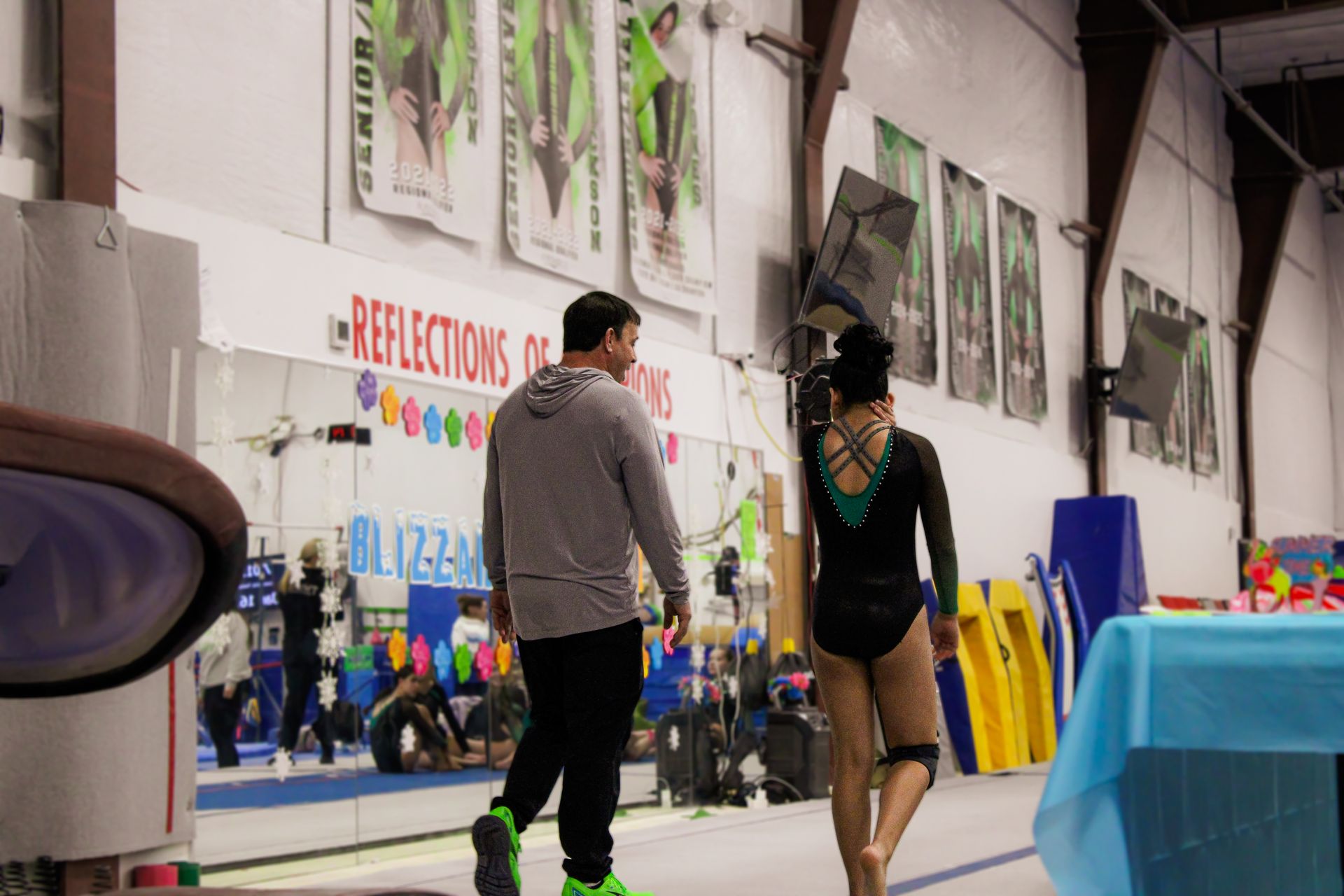 Two people walking in a dance studio, with mirrors, posters, and mats along the wall.