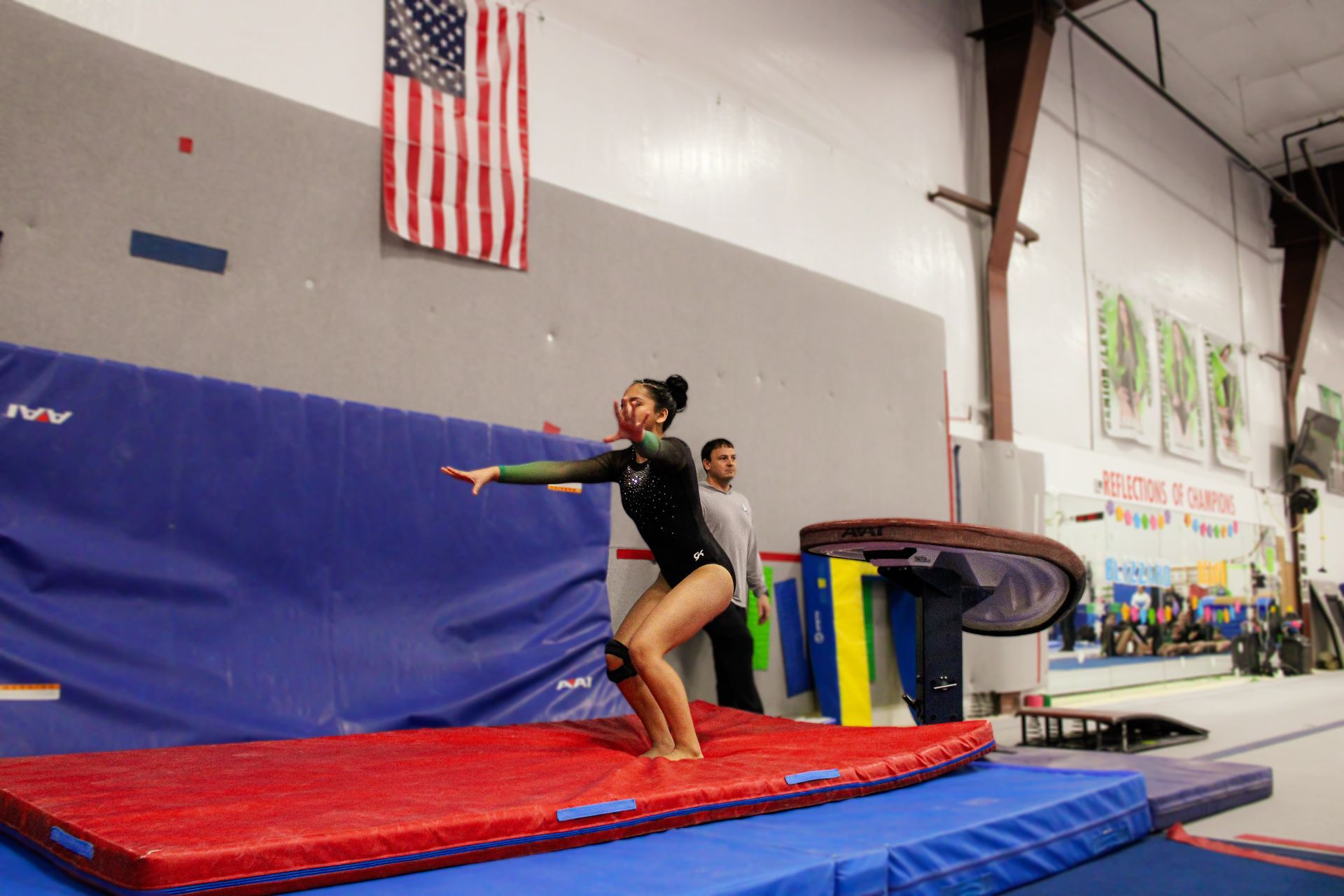 Gymnast balancing on a red mat in a gym, with a trampoline and U.S. flag in the background