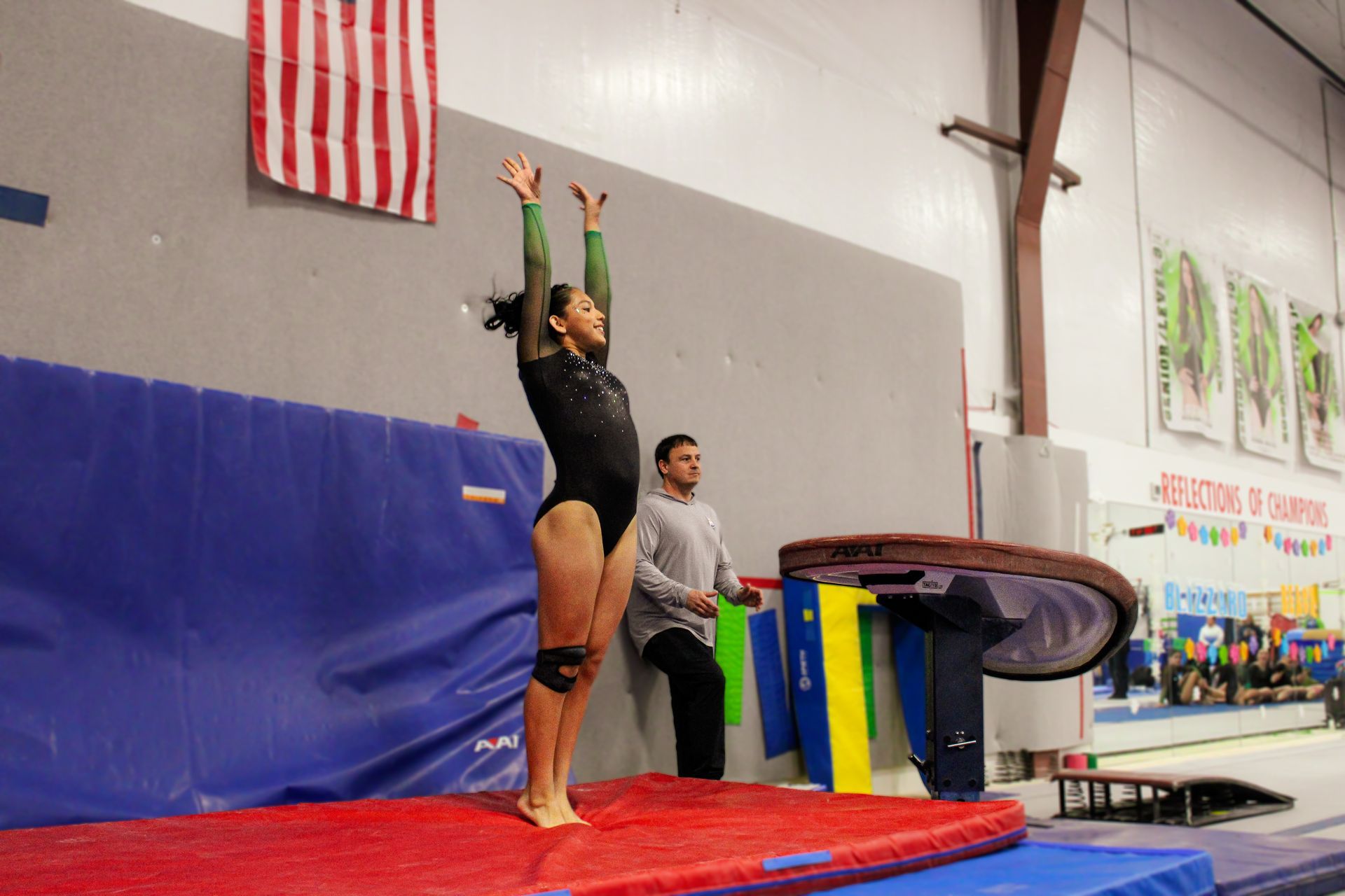 Gymnast in black leotard stands on mat with arms raised beside coach and uneven bars.