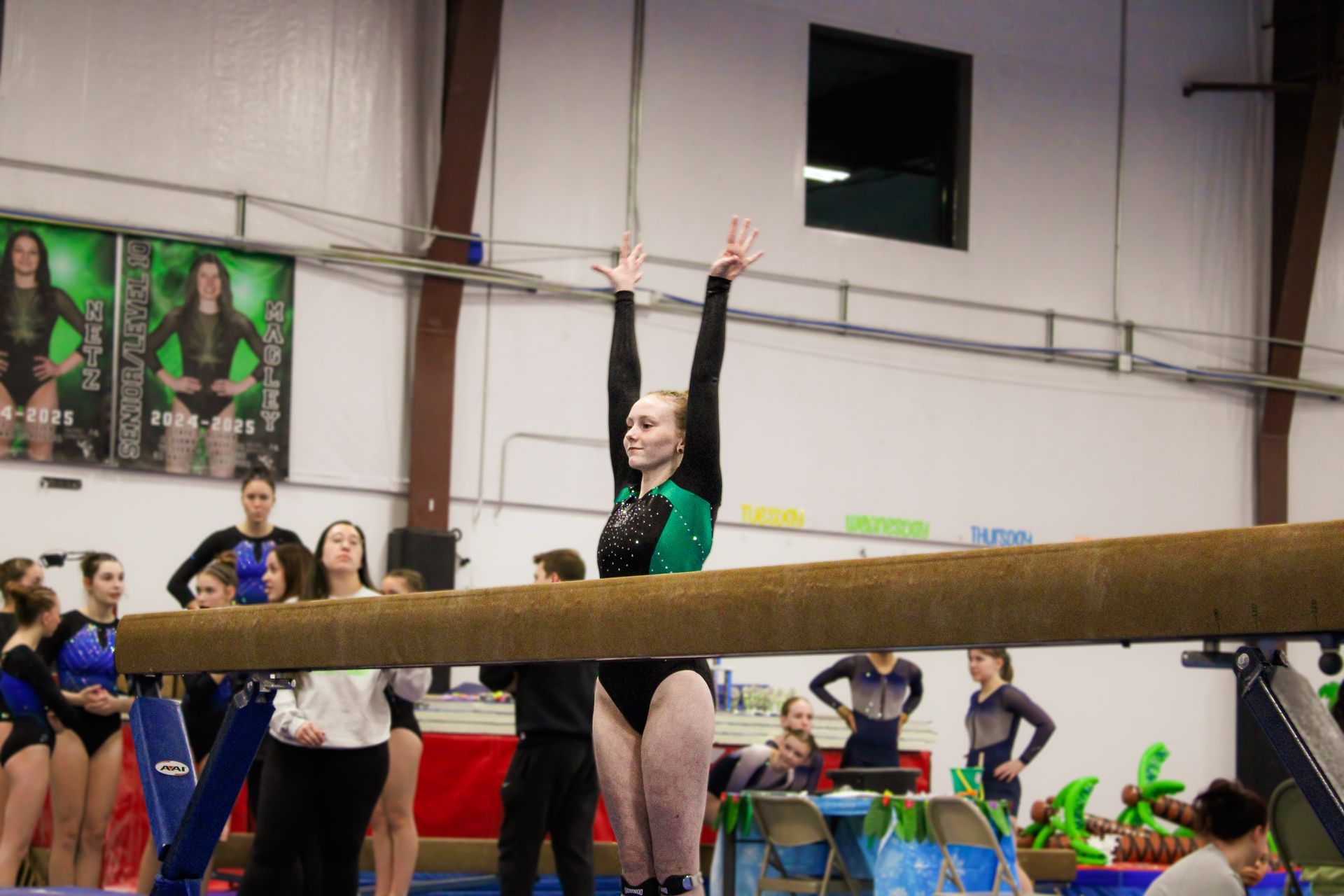 Gymnast balancing on a beam with arms raised in an indoor gym, teammates and posters in the background