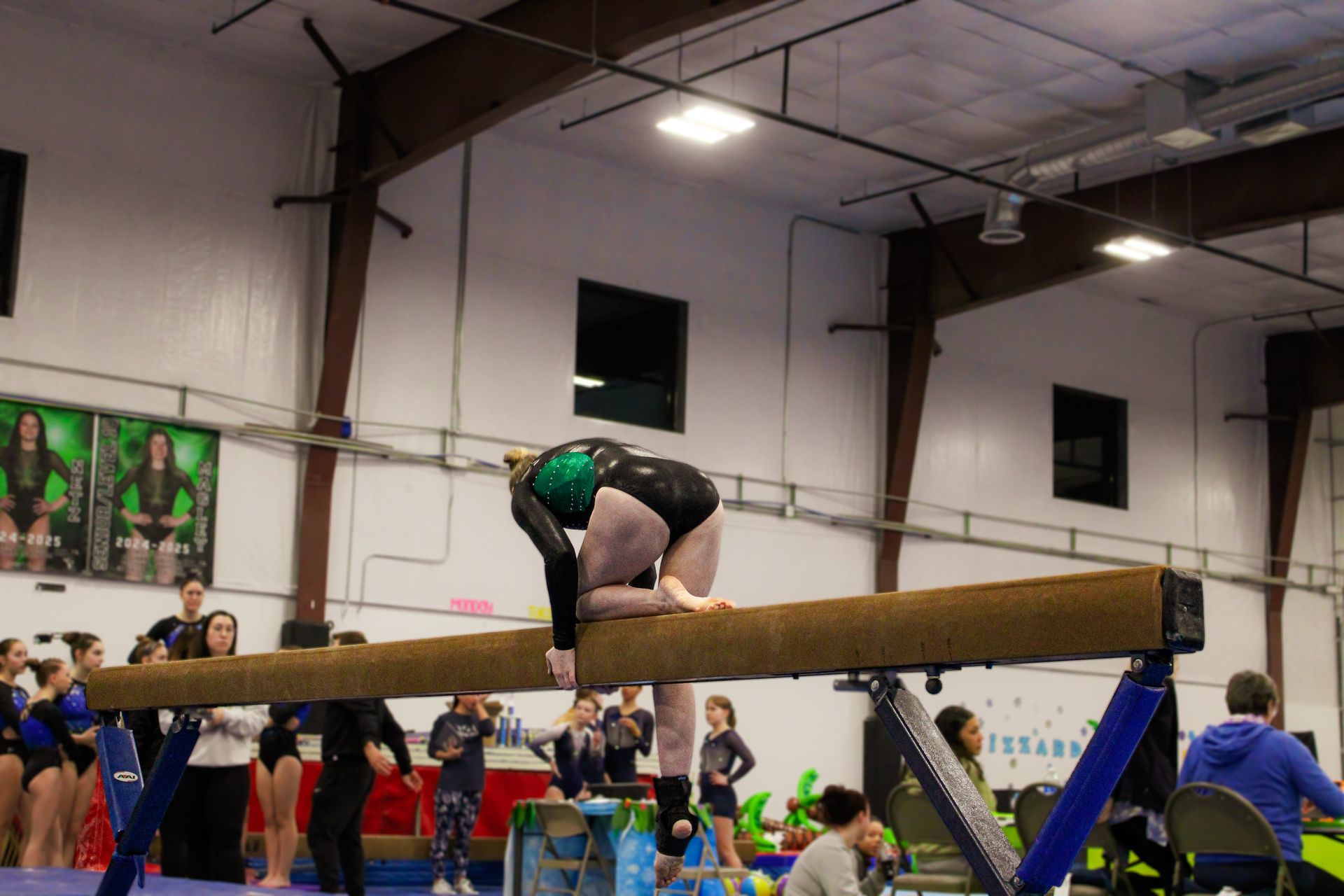 Gymnast balancing on a beam in an indoor competition gym, surrounded by spectators.