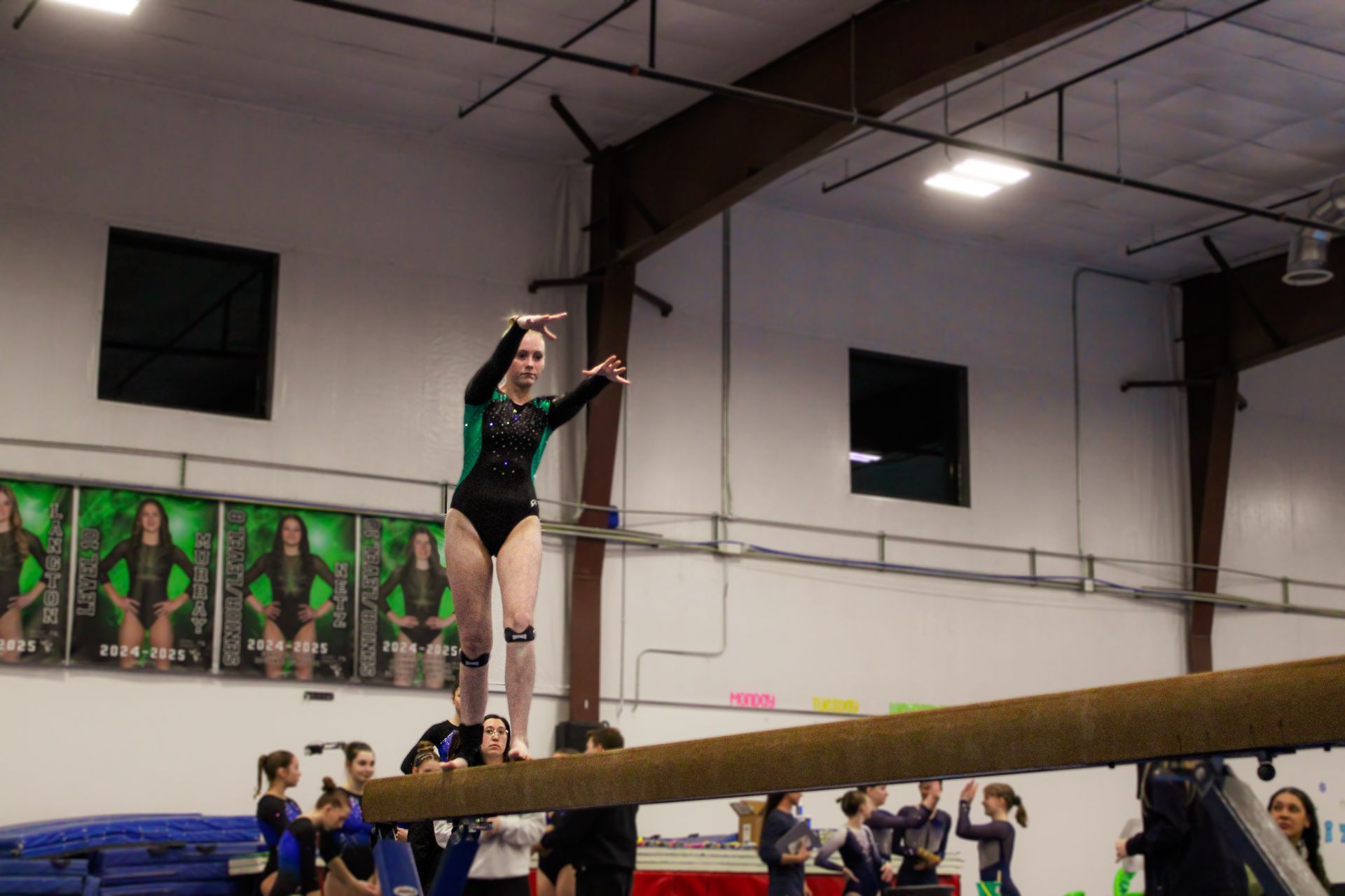 Gymnast balancing on a beam in an indoor gymnasium, arms raised for balance.