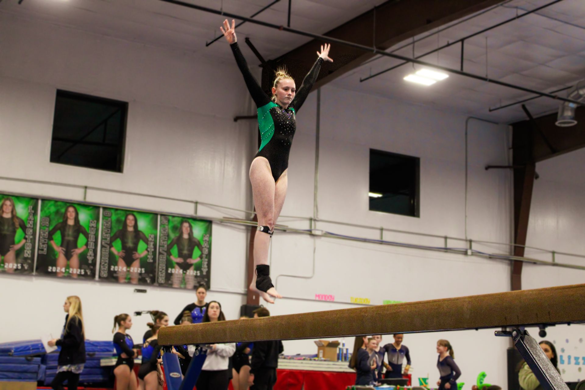 Gymnast balancing on a beam in a gym, wearing a green leotard with arms raised overhead