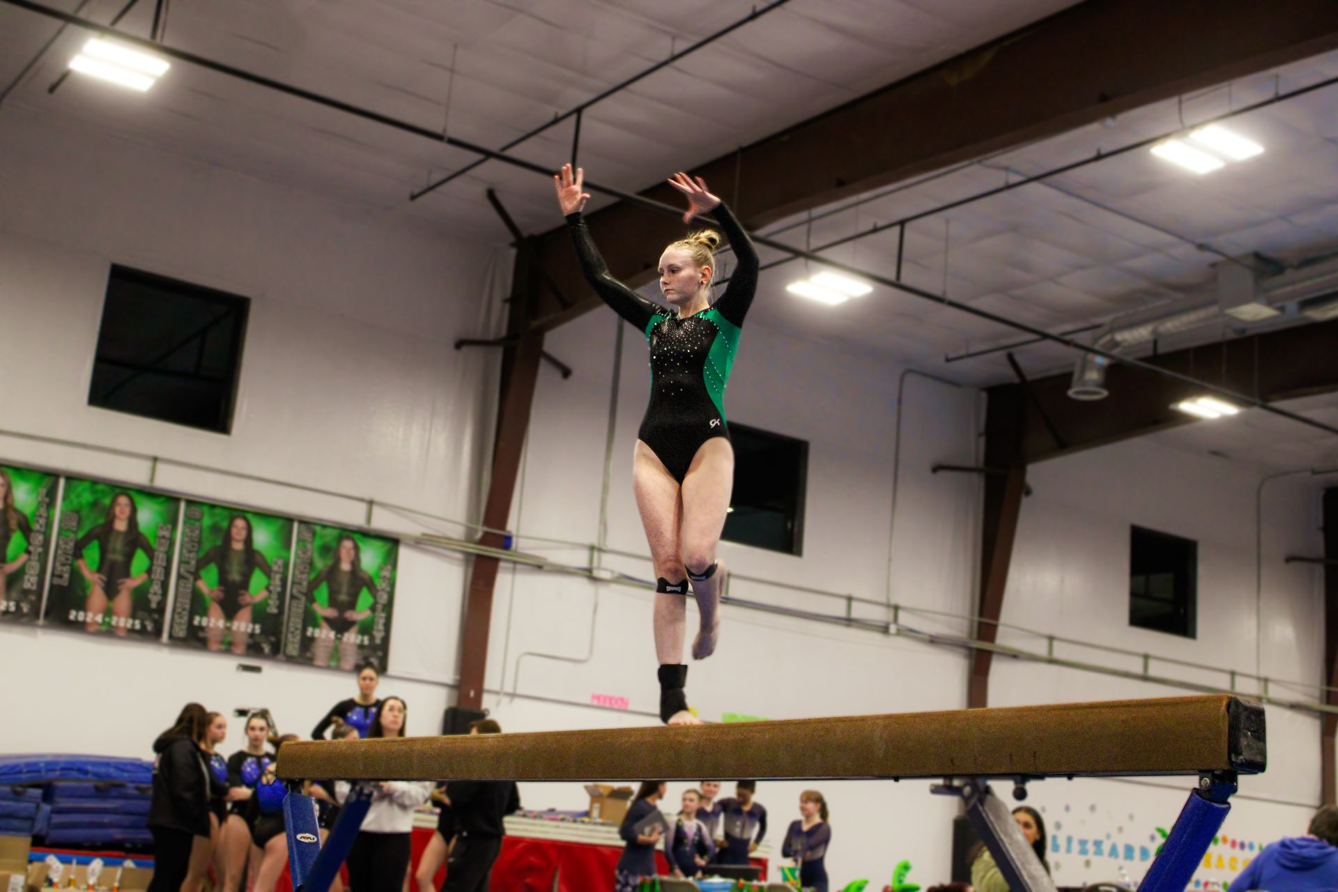 Gymnast balancing on a beam indoors with arms raised, wearing a black and green leotard.