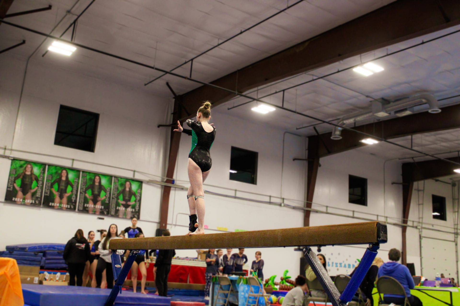 Gymnast balancing on a beam in an indoor gym competition, with spectators and mats below.