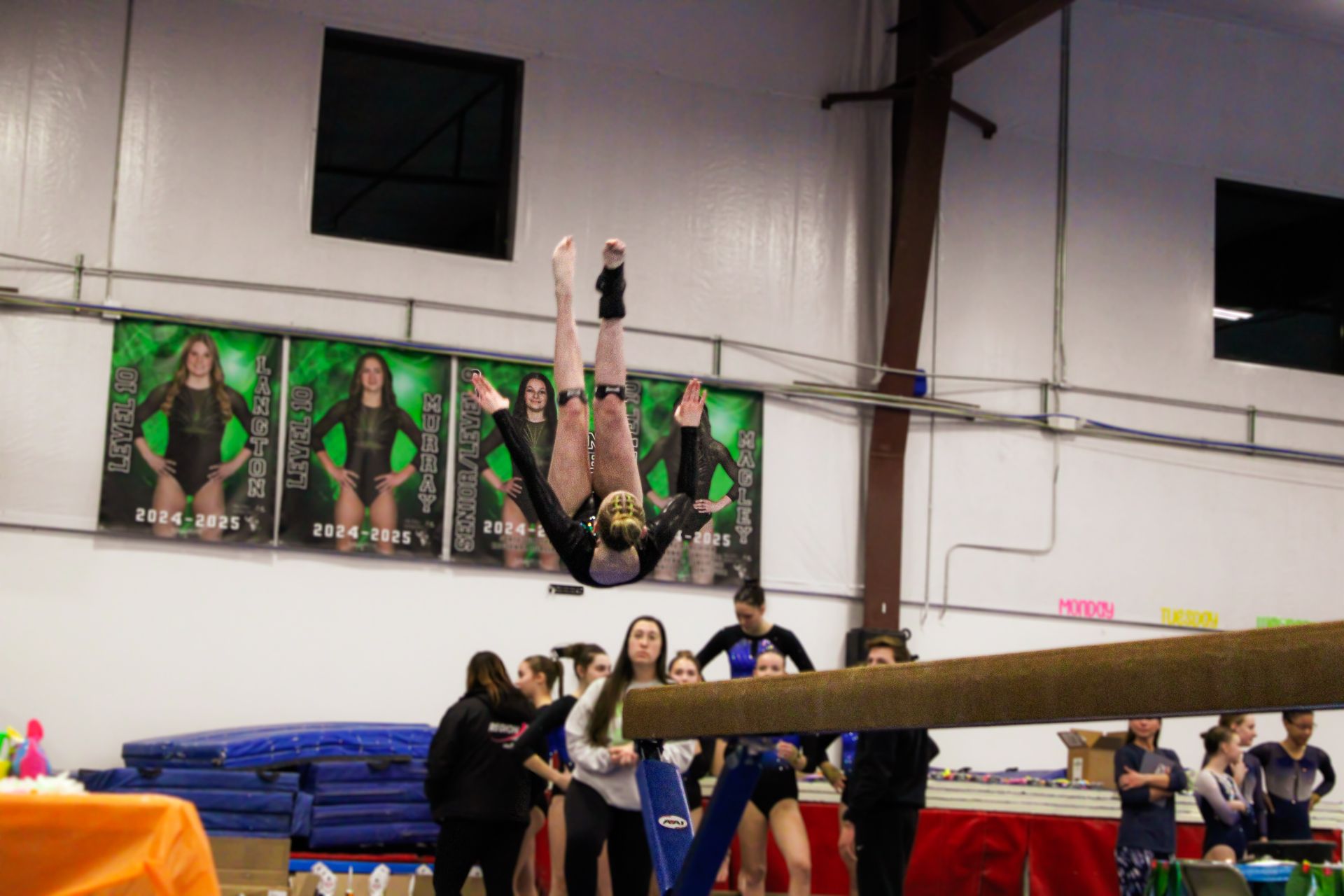 Gymnast performing a handstand on a balance beam in a gymnasium, with spectators and posters in the background