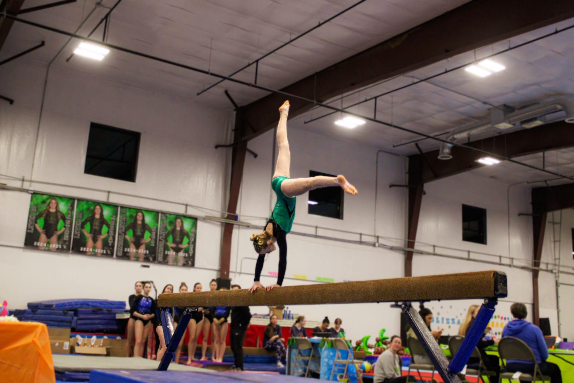 Gymnast balancing upside down on a beam in a gym, with teammates and spectators in the background