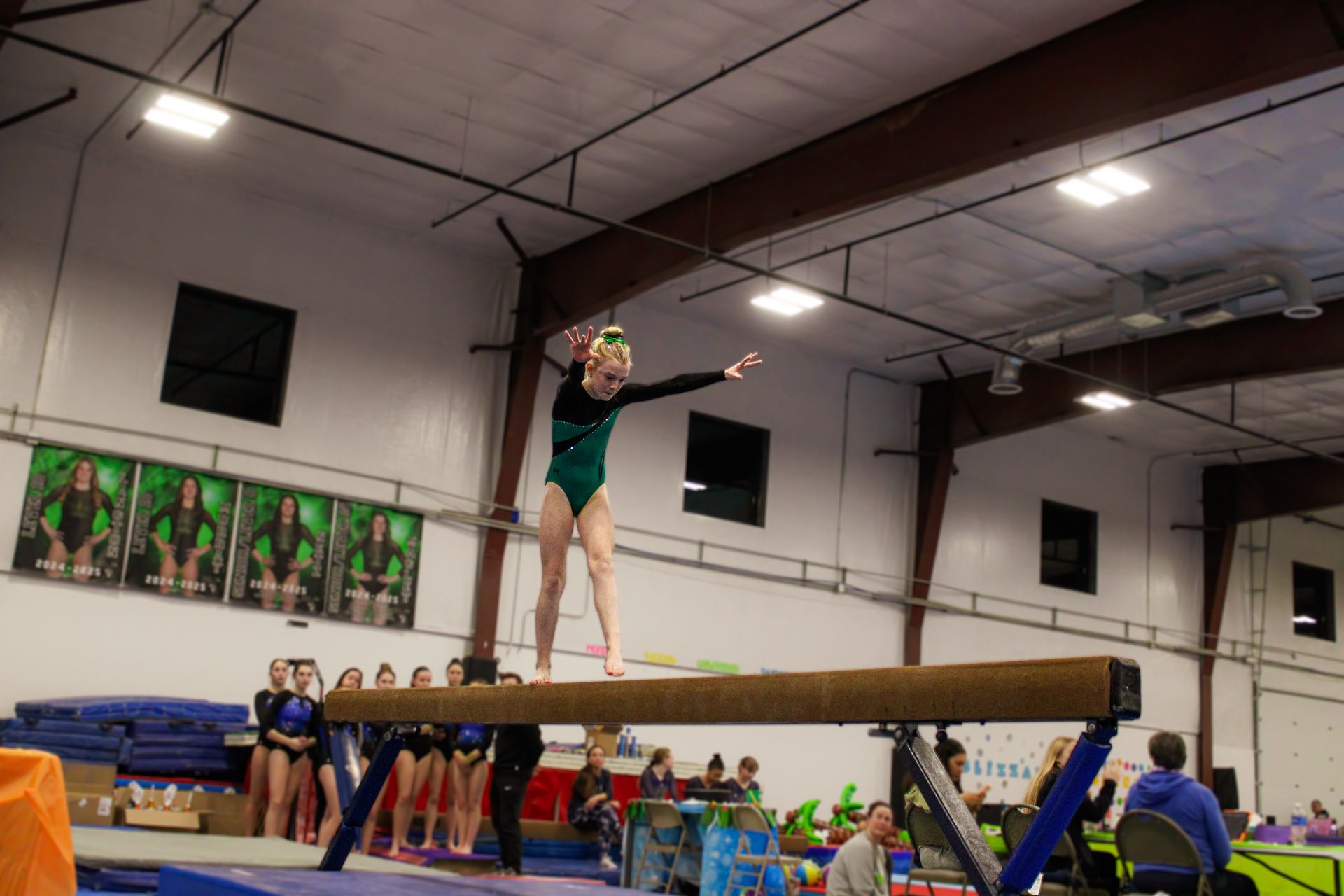 Gymnast balancing on a beam in a gymnasium, arms outstretched, with teammates watching nearby