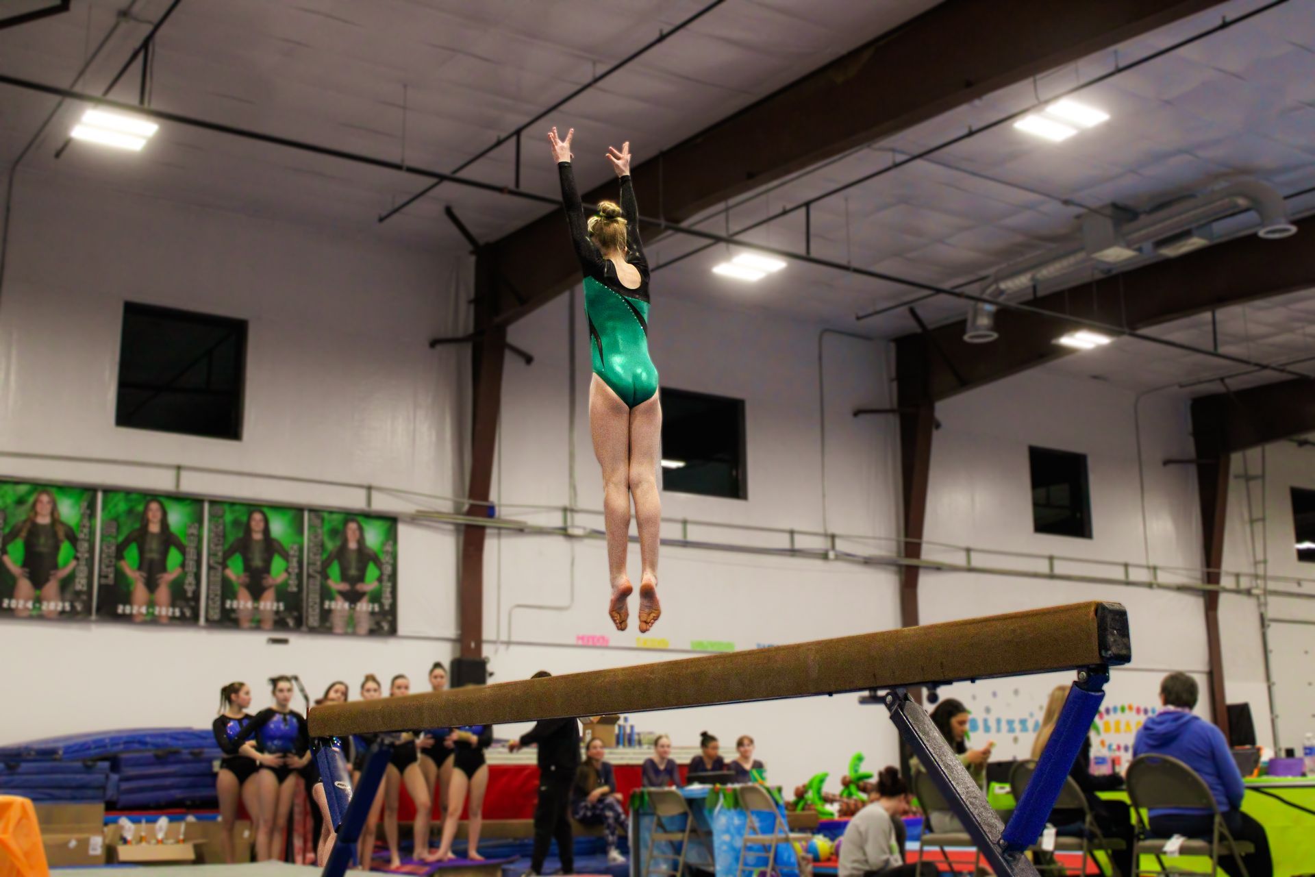 Gymnast in a green leotard performing on a balance beam in a gymnasium, with coaches and spectators nearby.