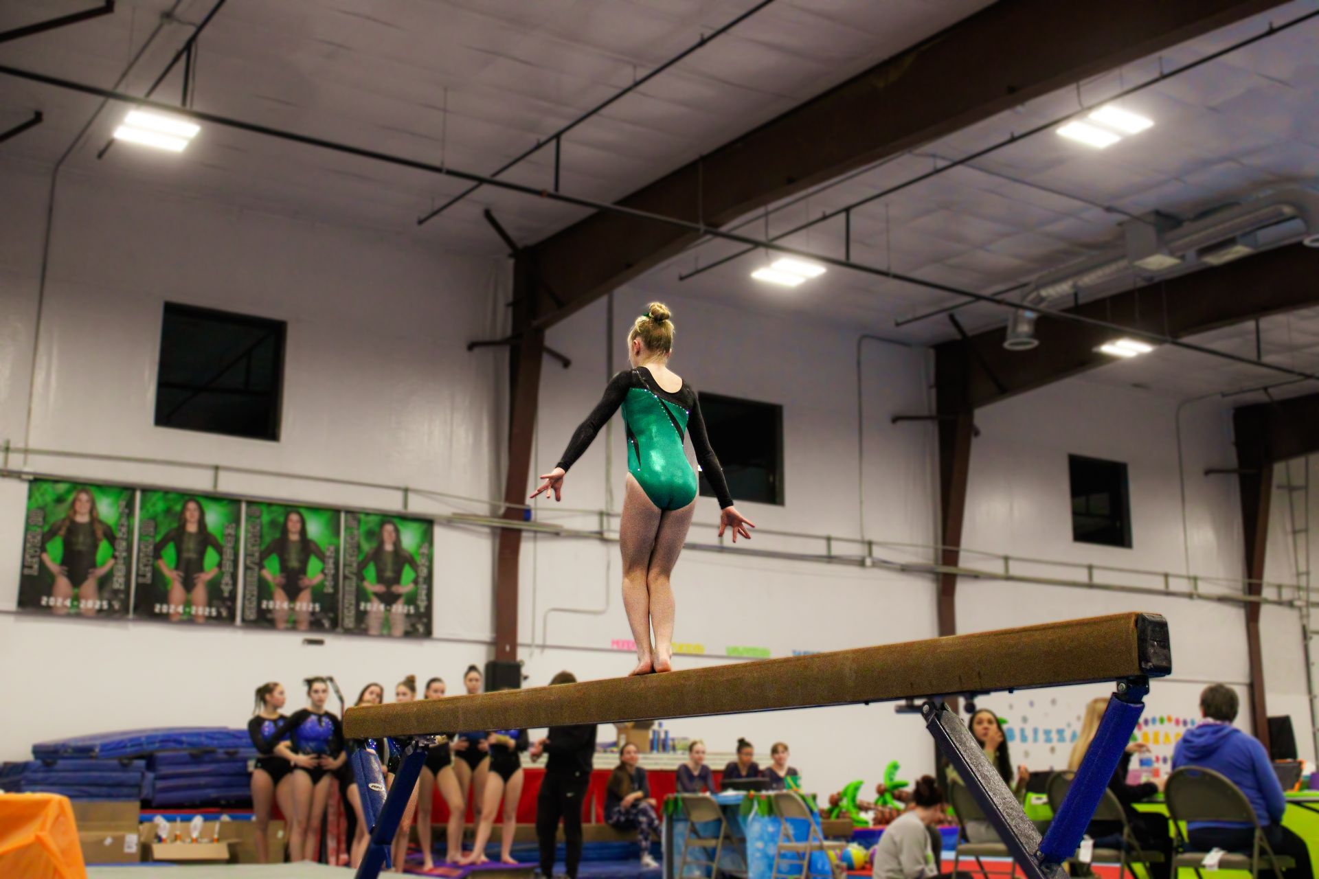 Gymnast balancing on a beam in a gym, with teammates and spectators in the background