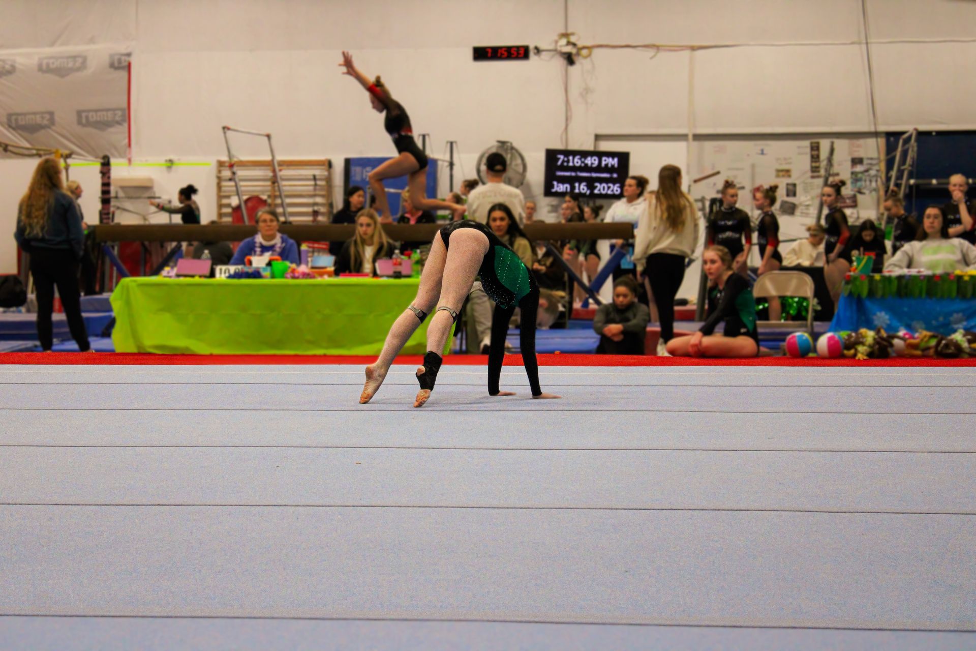 Gymnast in green balances on a blue mat during a floor routine, with judges and spectators in the background.