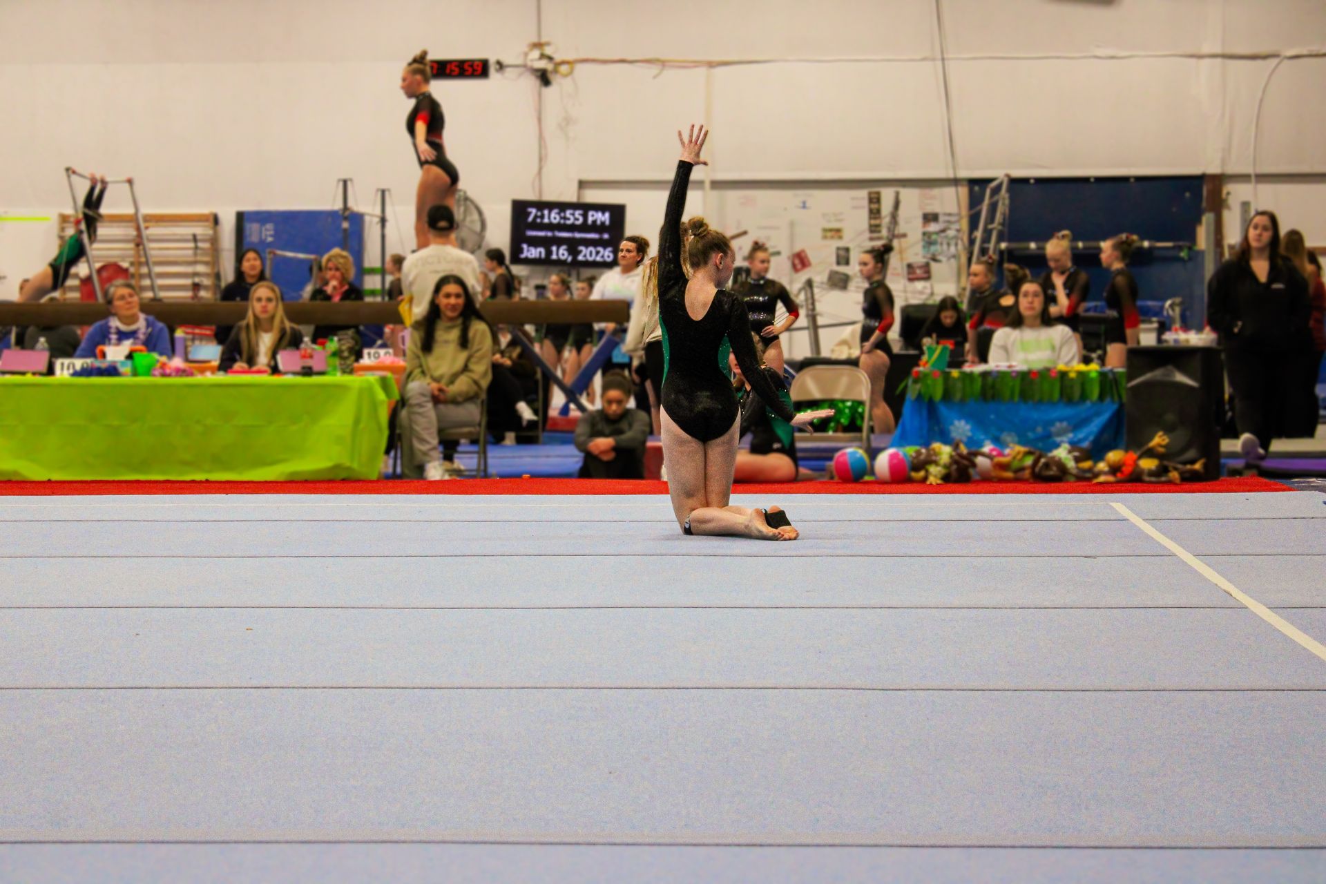 Gymnast performing a floor routine with one leg raised, in a gymnasium with spectators behind a barrier.