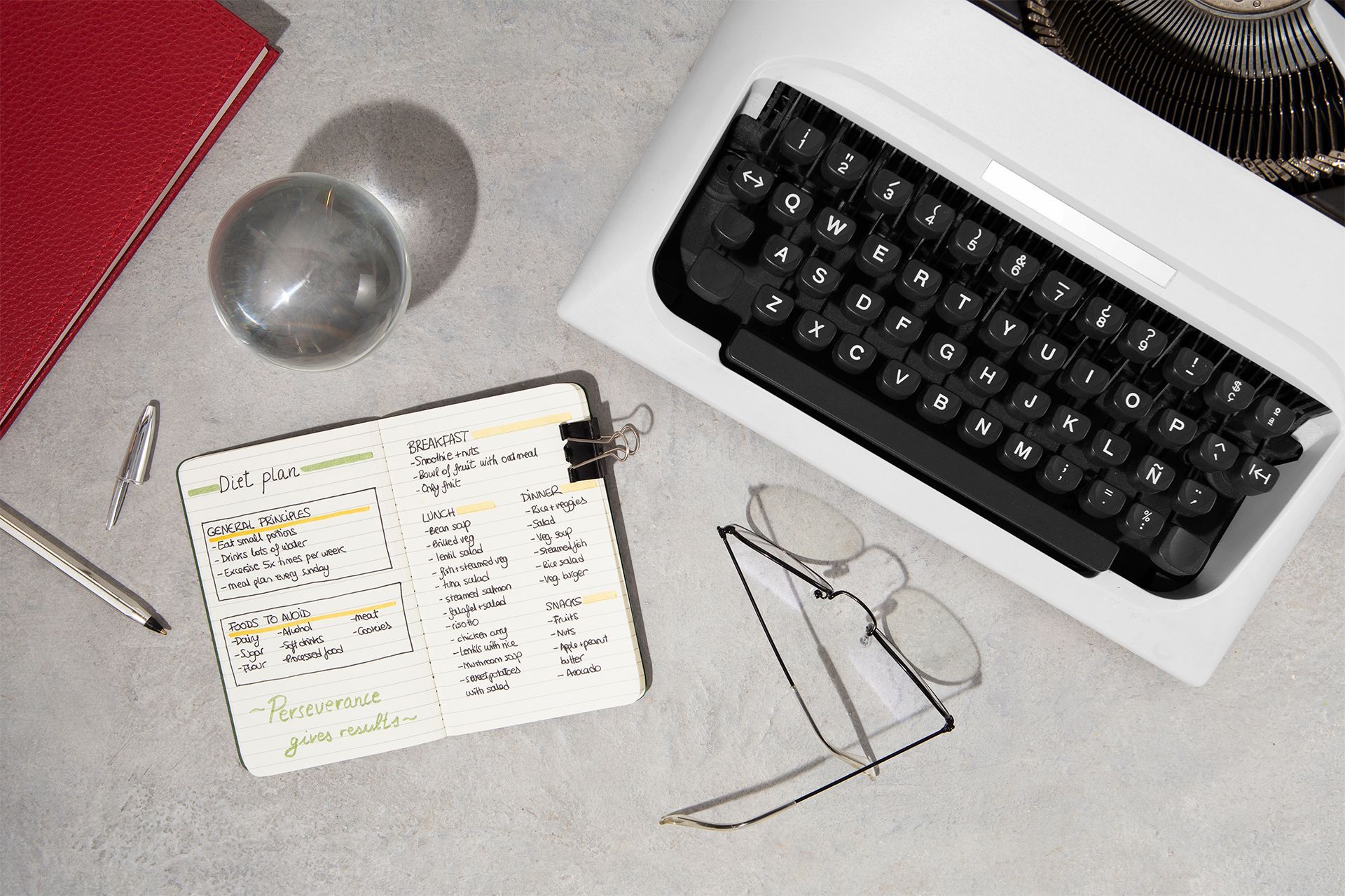 Desk with a typewriter, notebook, pen, red journal, and clear glass ball. Desk with a typewriter, notebook, pen, red journal, and clear glass ball.