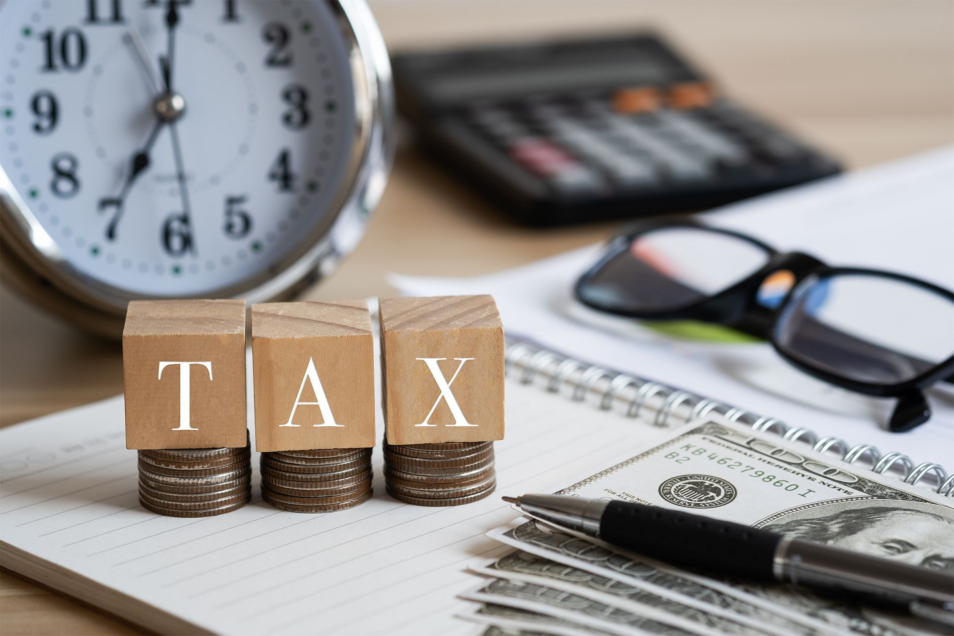 Tax-themed desk with wooden blocks, coins, calculator, glasses, money, and a clock. Tax-themed desk with wooden blocks, coins, calculator, glasses, money, and a clock.