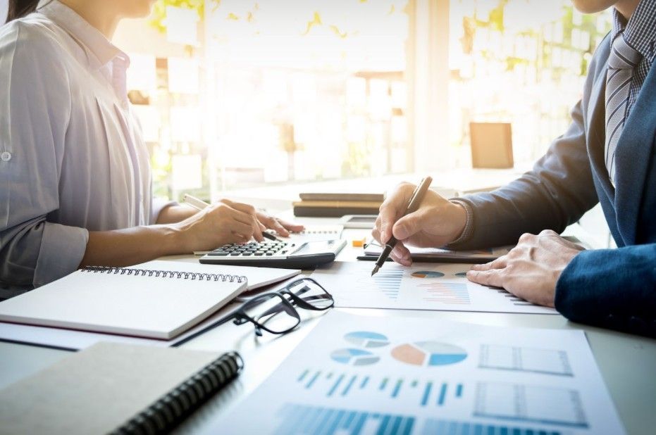 Two professionals review documents, charts, and a calculator at a desk in a bright, sunlit office.