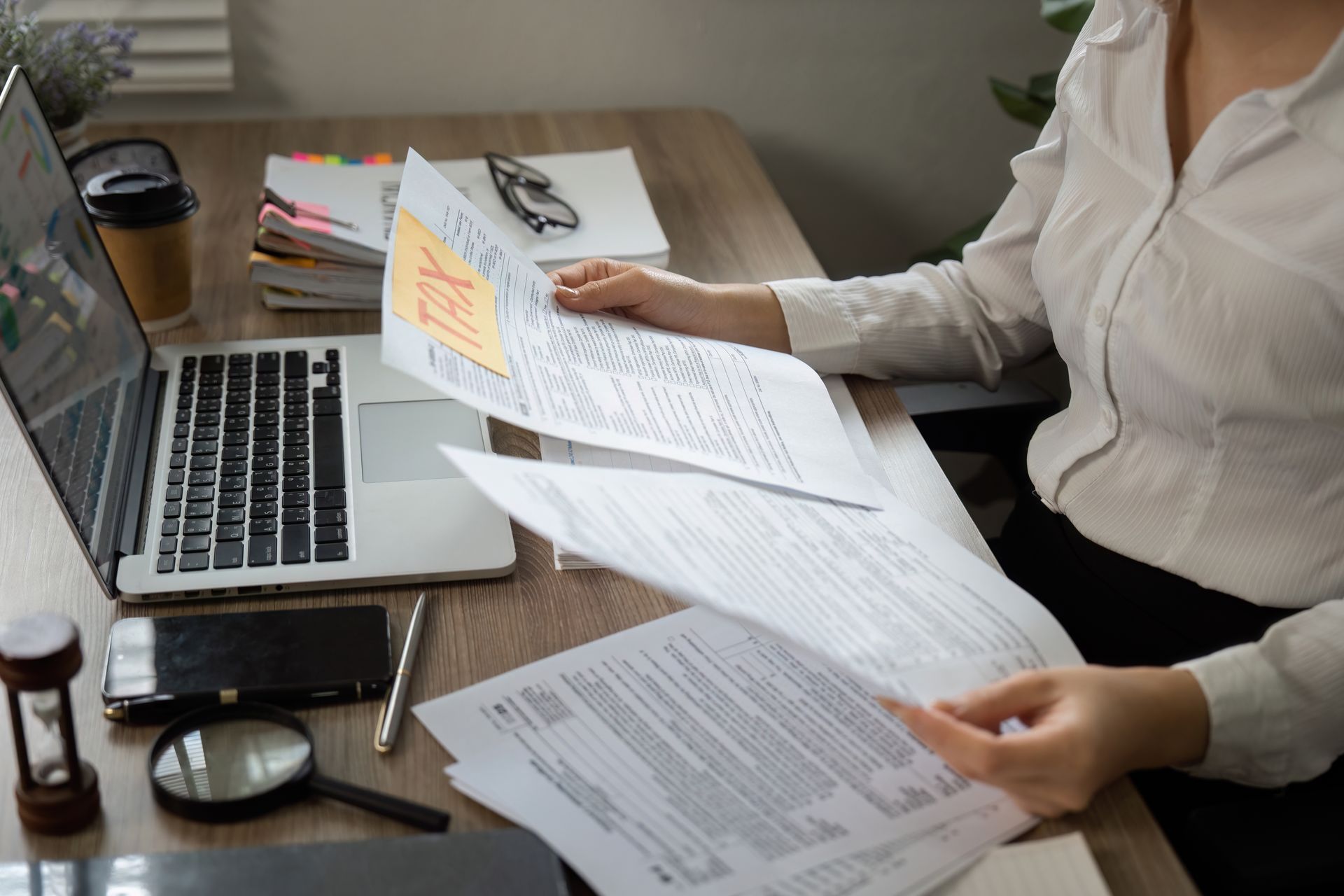 Person at desk reviewing papers beside an open laptop and coffee mug