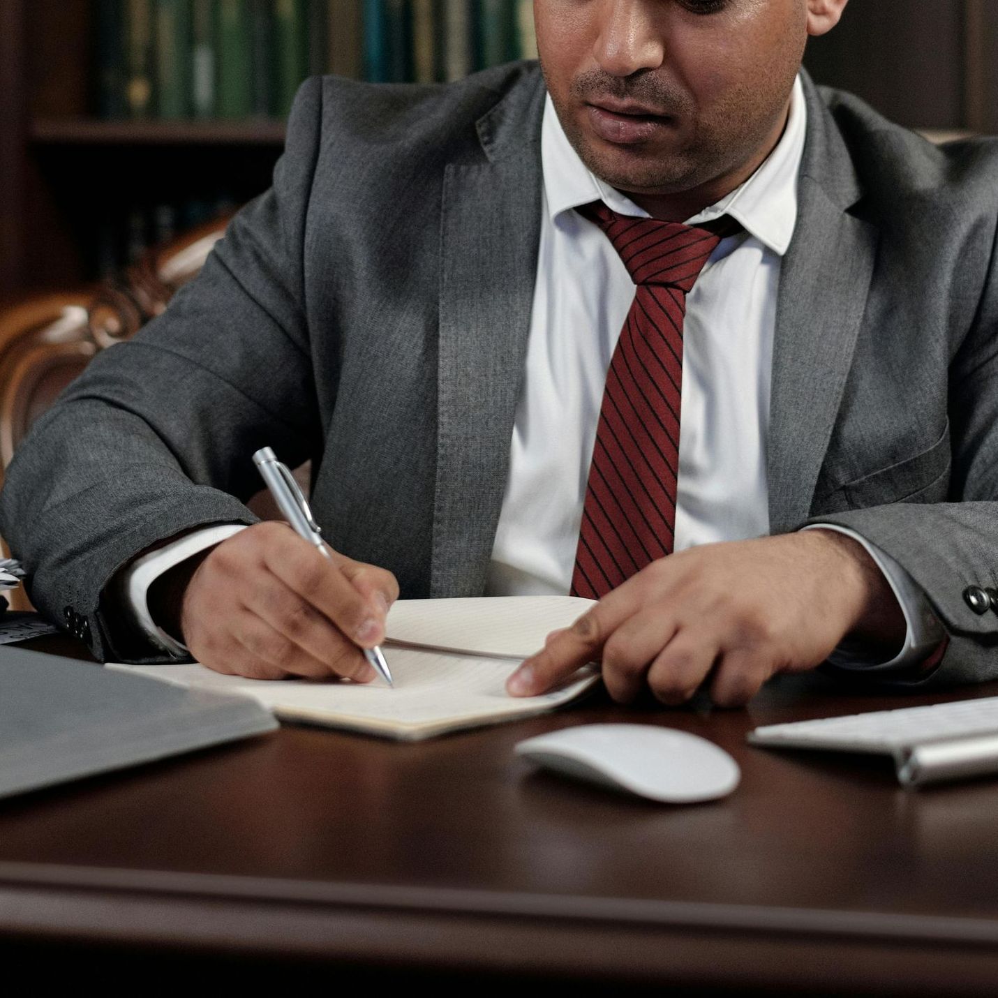 A person in a gray suit and red tie sits at a desk, writing in a notebook near a computer mouse and keyboard.