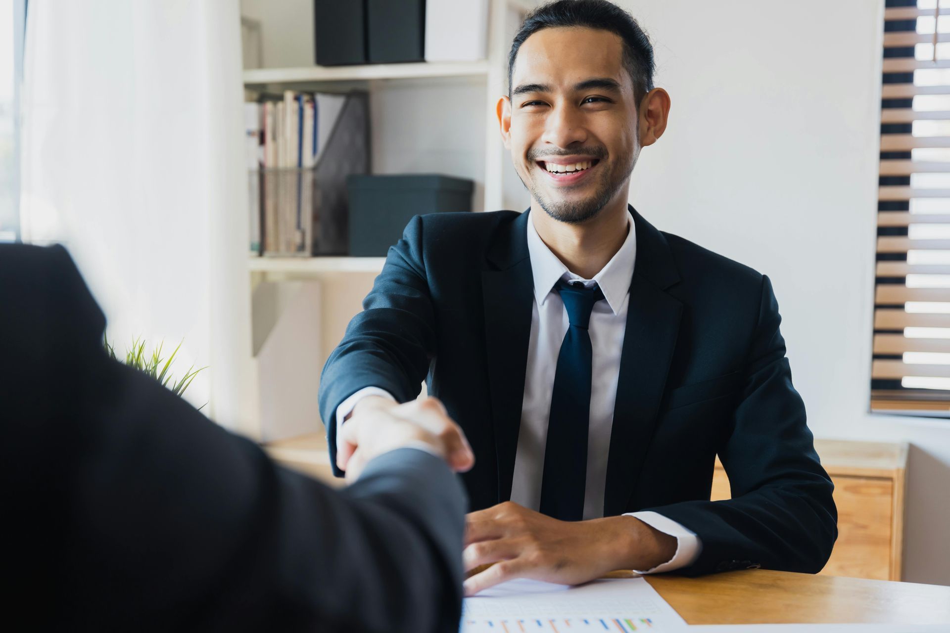 A professional in a suit smiles and shakes hands with a colleague across a desk in a well-lit office.