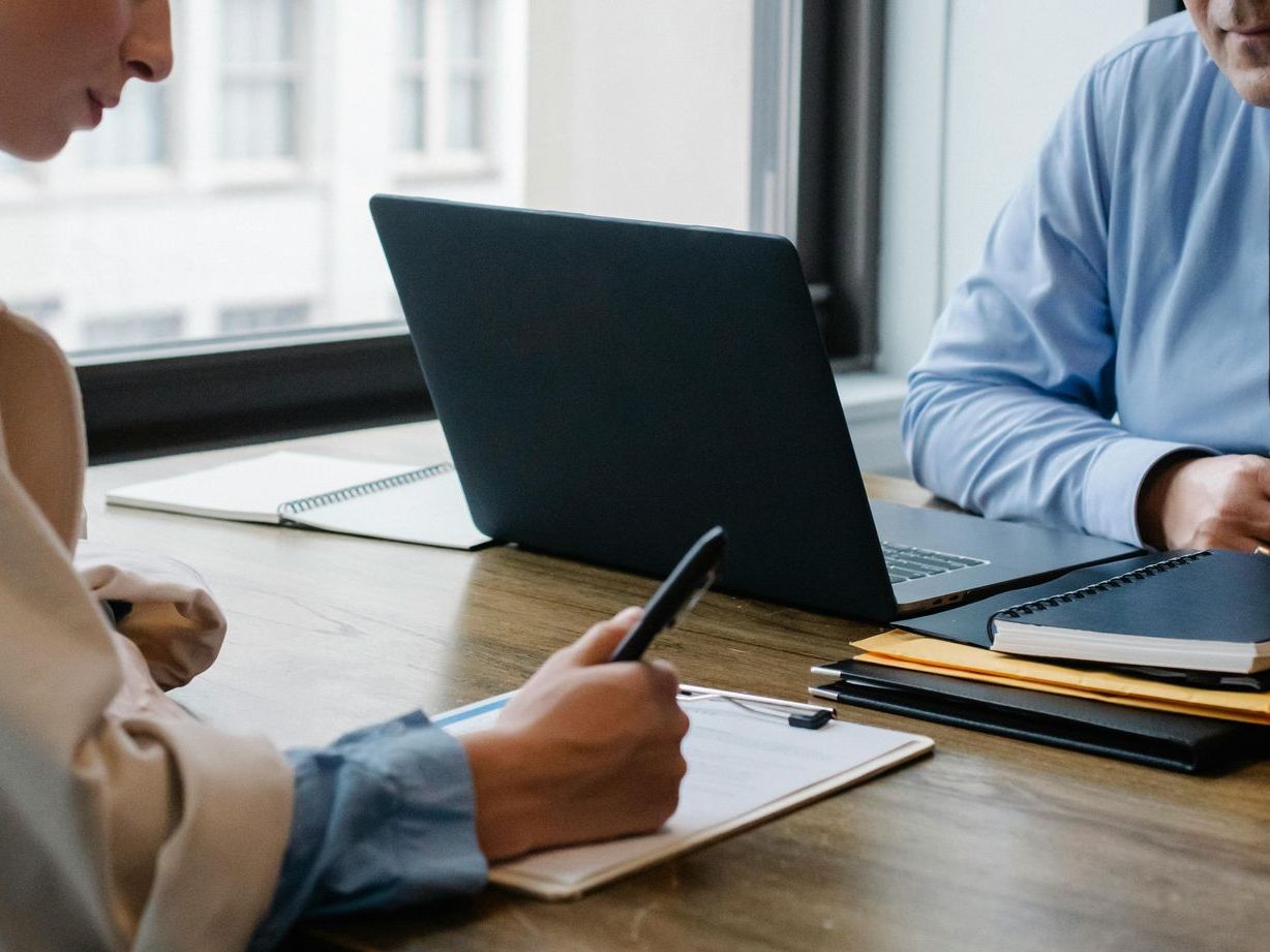 Two people at a desk with a laptop and documents; one person writes on a notepad while the other sits nearby.
